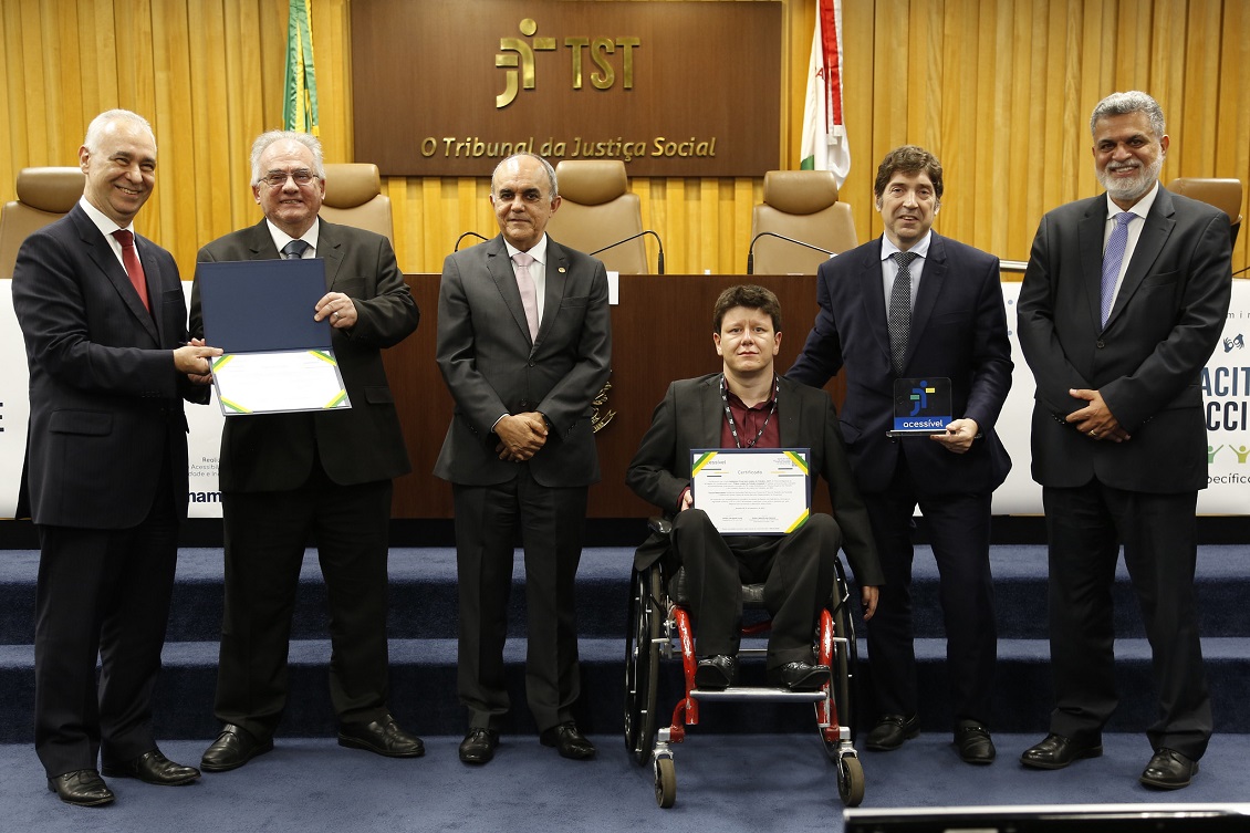 Servidor Sisenando ao centro, na cadeira de rodas, segurando certificado ao lado do juiz Guilherme Feliciano com o prêmio nas mãos. Ainda na foto, dirigentes do TST