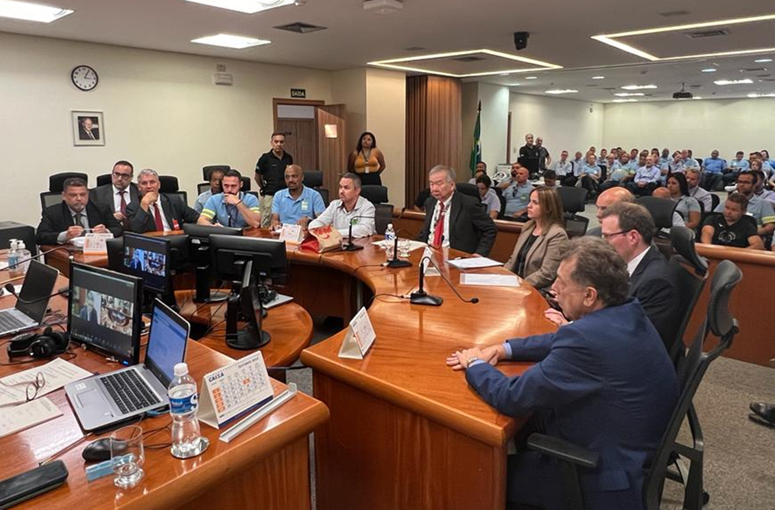 #ParaTodosVerem: sala de audiência com representantes do sindicato à esquerda da imagem, e da empresa, à direita, em frente a computadores em mesa semicircular. Ao fundo o auditório está lotado de trabalhadores uniformizados com camiseta azul clara. 