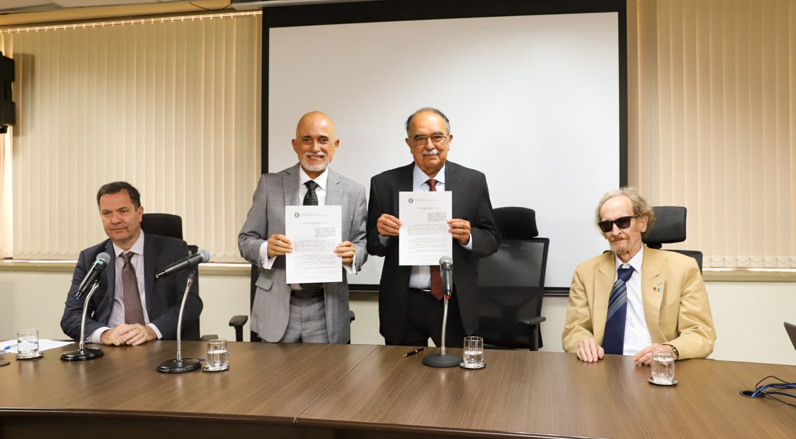 Doutor Samuel e doutor José Jozefran, atrás de uma mesa, em pé, mostram o Acordo de Cooperação que assinaram. Sentados, à mesa, estão mais dois homens.