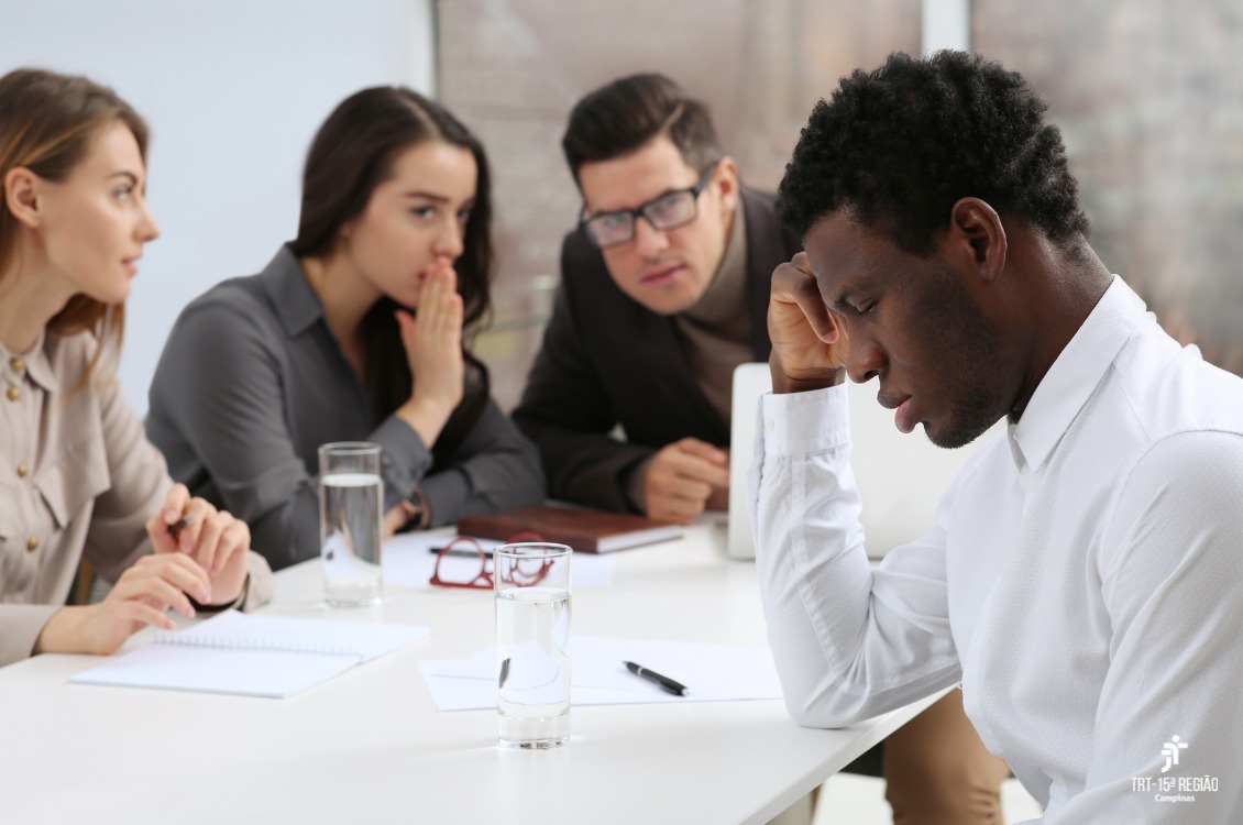 Sentados à mesa de trabalho, um homem negro é ridicularizado por três colegas