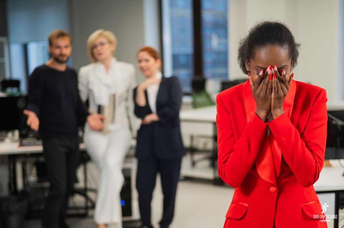 mulher negra está com mãos postas escondendo o rosto. Ao fundo, três pessoas conversam de forma escusa.