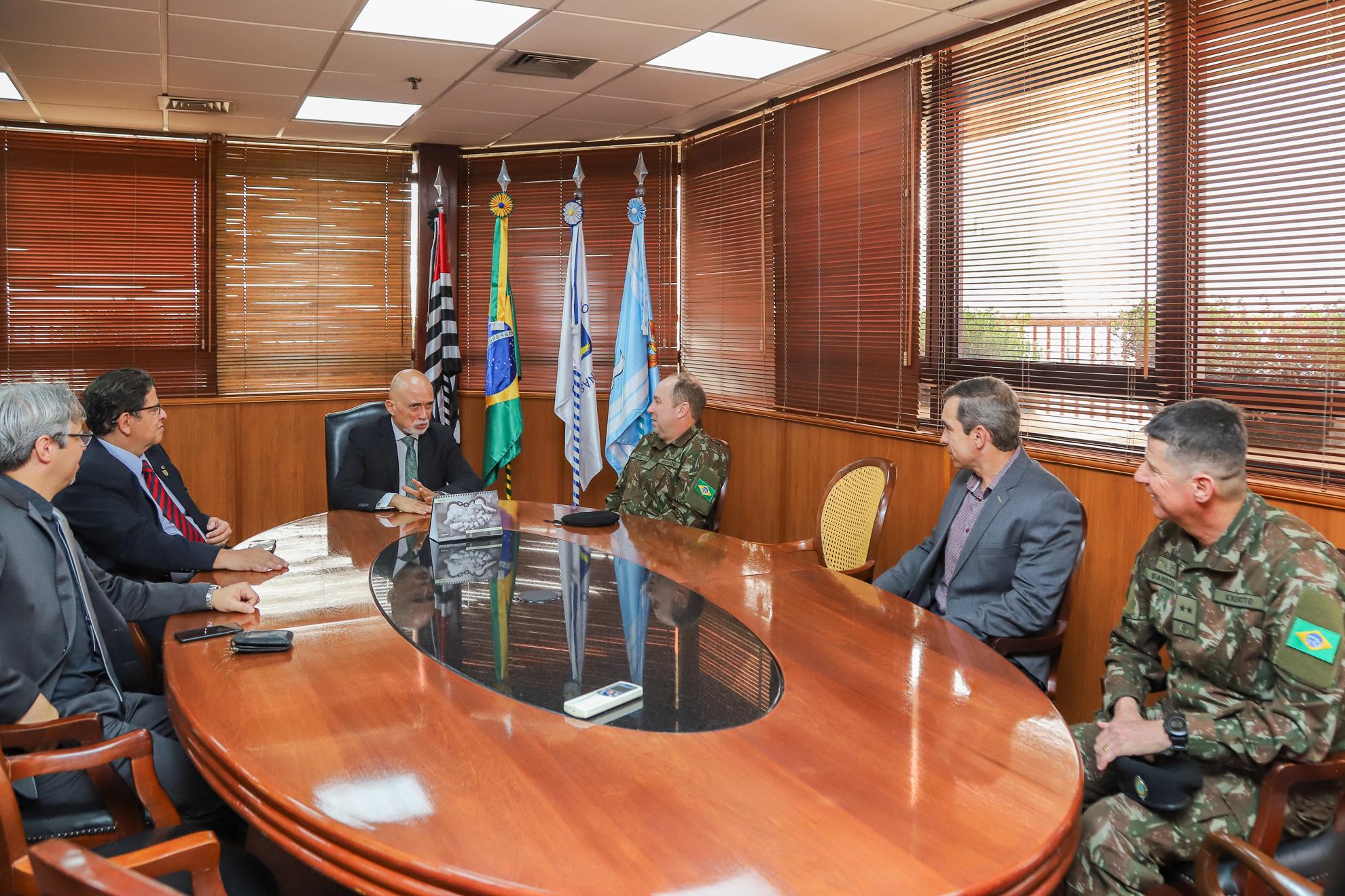 A foto retrata uma mesa oval. Seis homens sentados