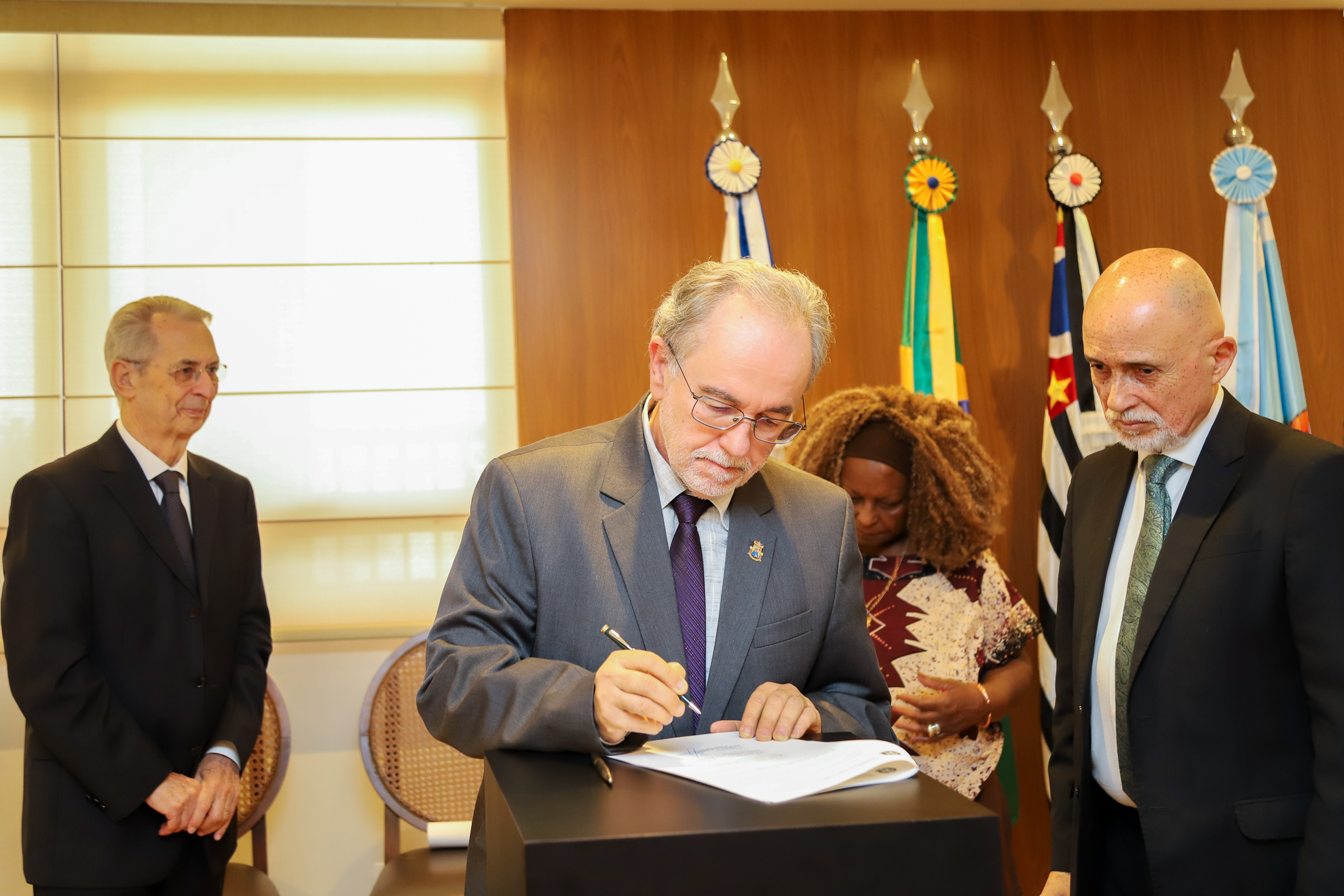 Reitor da PUC assina o termo, sendo observado por membros da universidade e pelo Dr. Samuel