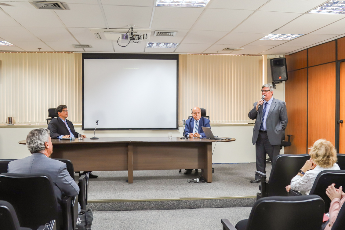 componentes da mesa alta, plateia, telão ao fundo e o desembargador João Alberto está de pé, falando ao microfone