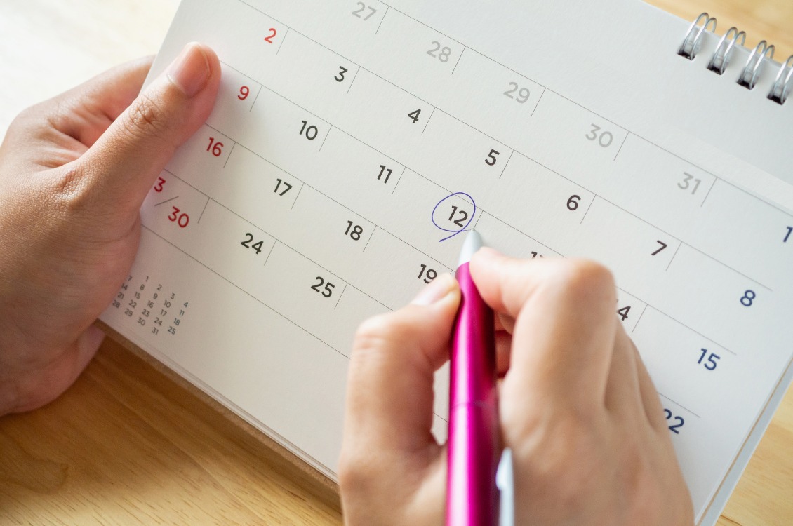  A imagem mostra uma mão segurando um calendário enquanto a outra mão marca o dia 12 com uma caneta rosa. O calendário é sobre uma superfície de madeira clara.