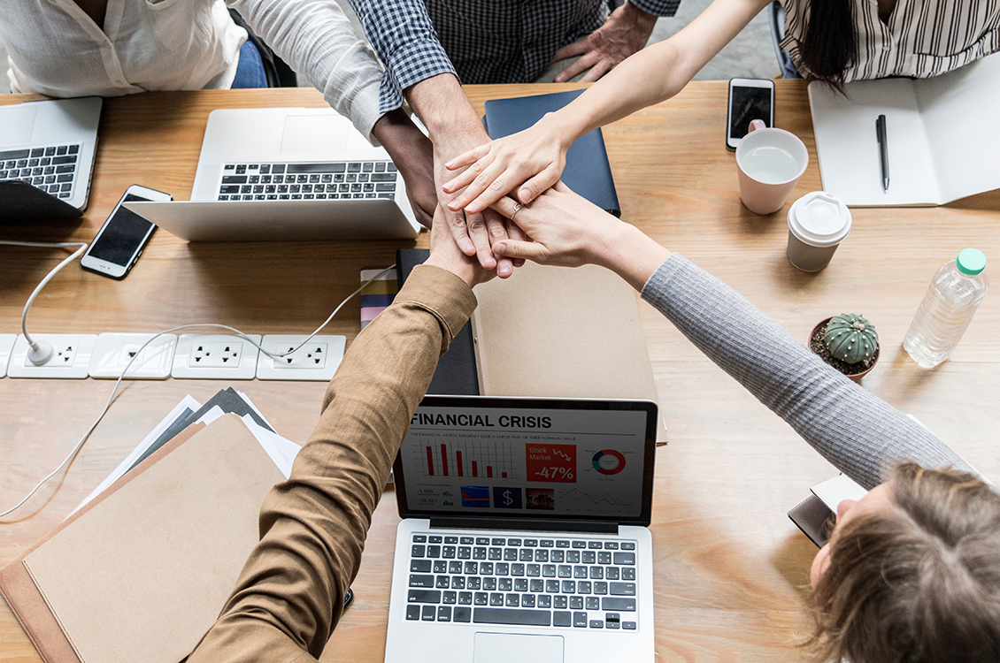 imagem mostra trabalhando em equipe com mãos entrelaçadas e computadores em cima da mesa