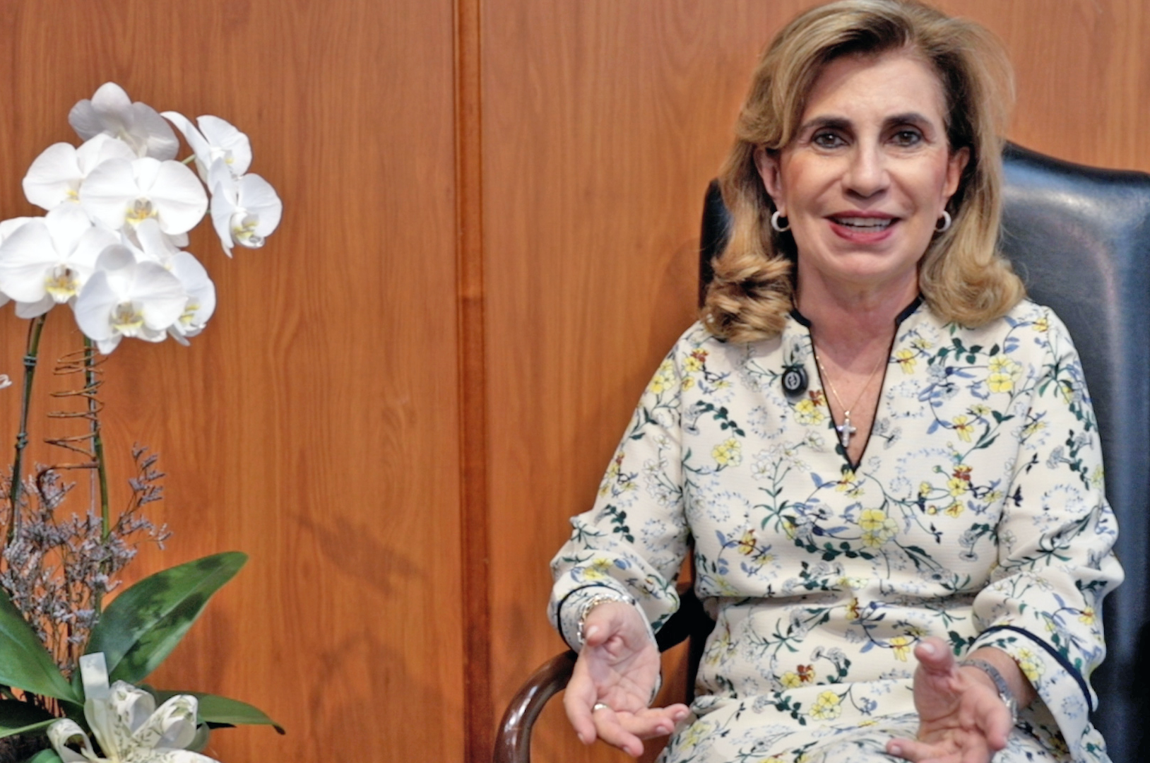 imagem com fundo de madeira. Na lateral direita da tela, Dra. Ana Paula está sentada, sorrindo e gesticulando. Na lateral esquerda da tela, uma orquídea branca