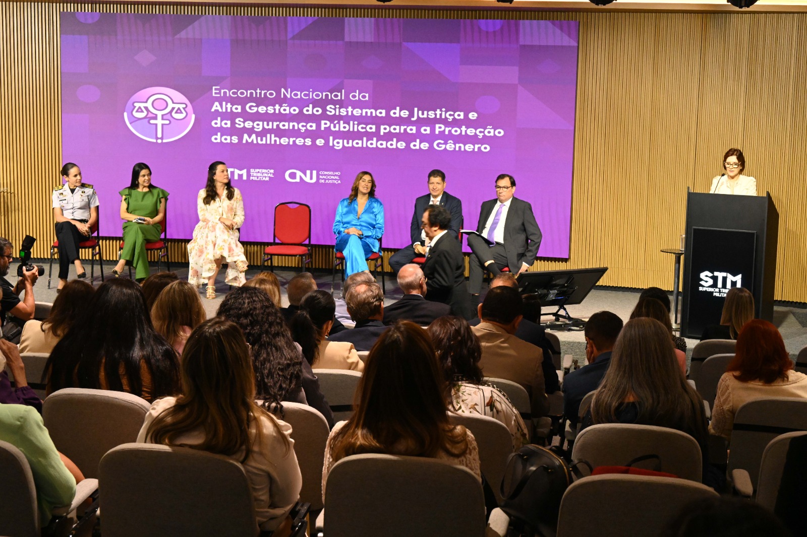 seis componente da mesa alta estão sentados em cadeiras dispostas no palco. Uma mulher fala ao microfone no púlpito, sendo observada pelo público.