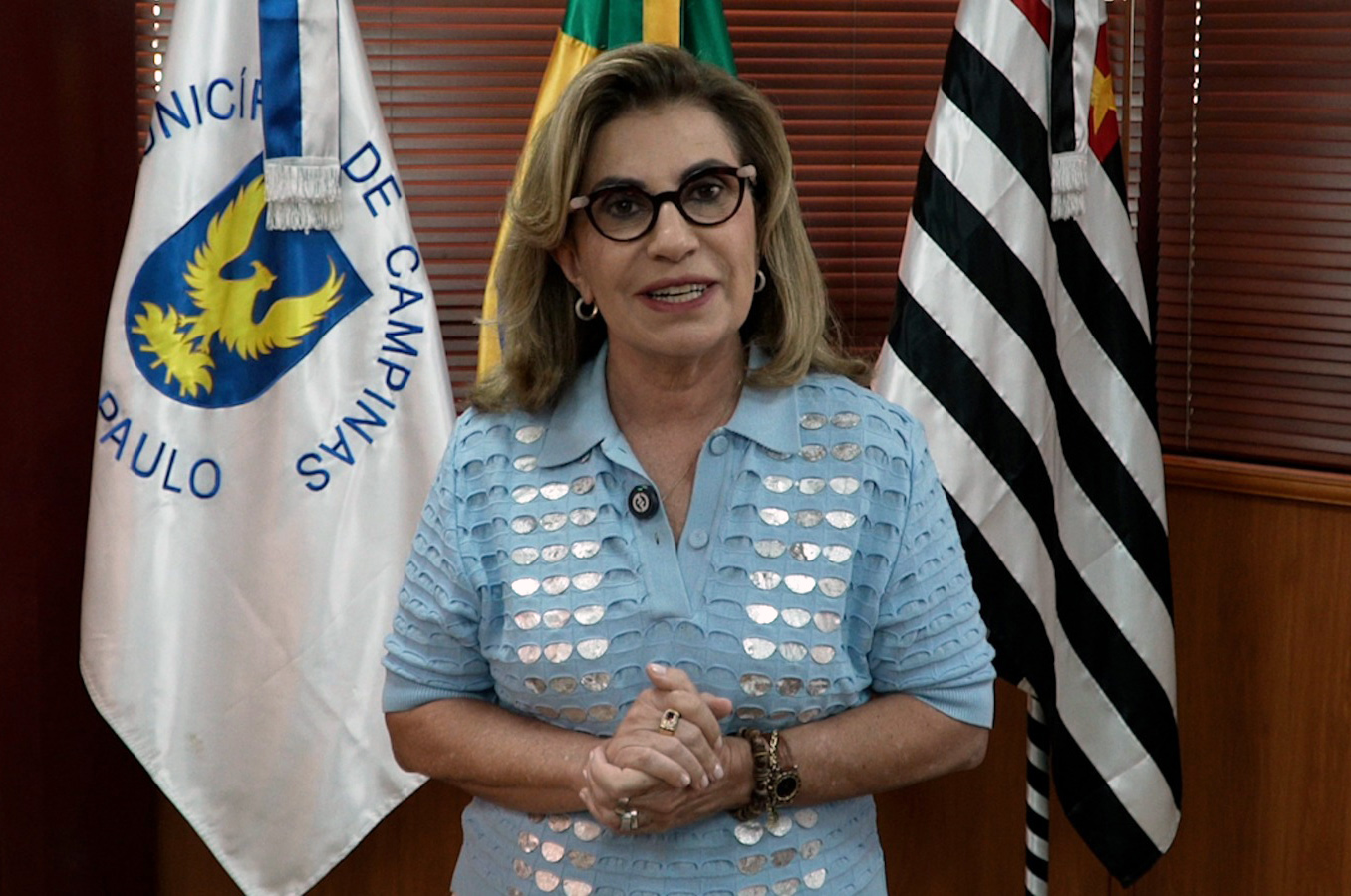 Dra. Ana Paula Lockmann está de pé em frente a três bandeiras, de blusa azul clara e mãos juntas na altura da cintura