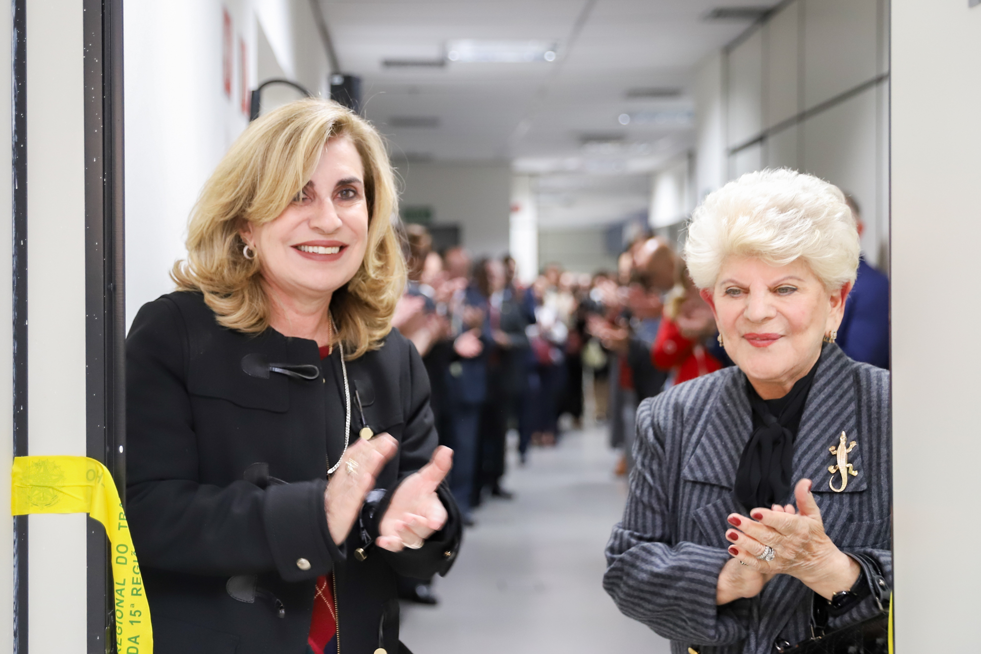 Dra. Ana Paula e Dra. Eliana aplaudem após desenlaçar a fita amarela de inauguração, sendo observadas pelo público ao fundo.