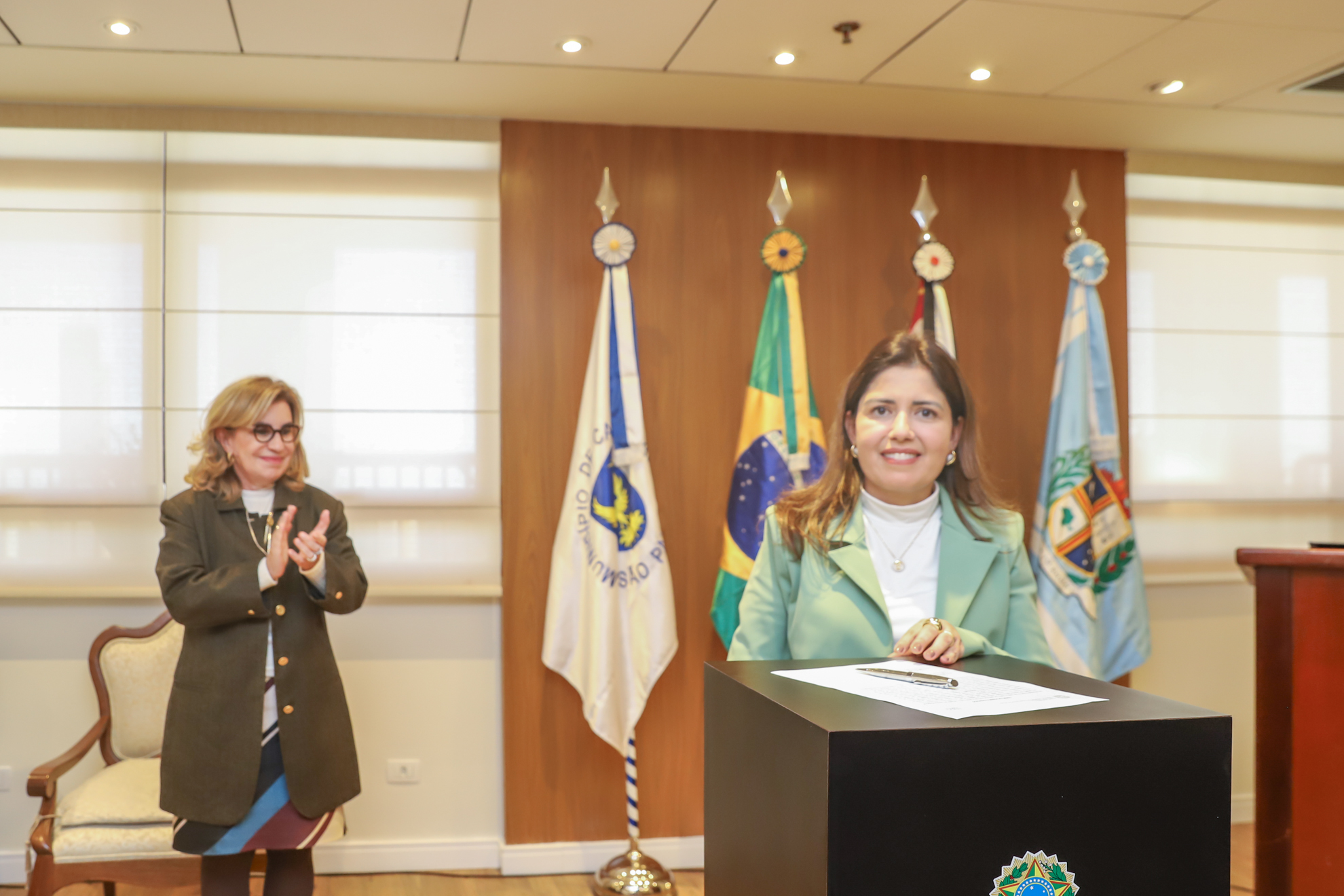 juíza está atrás de dispositivo, olhando para câmera após assinatura de termo de posse. À esquerda da tela, Dra. Ana Paula aplaude o ato.