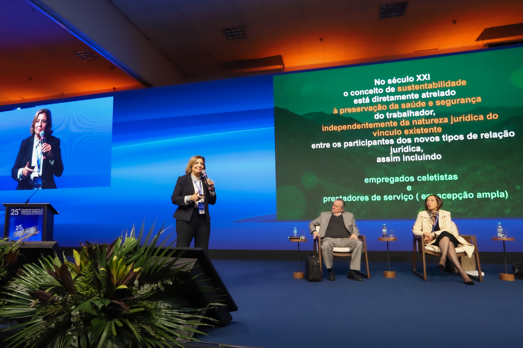 A imagem mostra a desembargadora do TRT-15 Tereza Aparecida Asta Gemignani em pé, falando ao público durante o 2º painel do 25º Congresso Nacional de Direito do Trabalho e Processual do Trabalho. Ela está ao lado de um telão que exibe o texto de sua apresentação sobre sustentabilidade e proteção à saúde e segurança do trabalhador no século XXI. Sentados no palco, o advogado Celso Antonio Pacheco Fiorillo e a desembargadora Erodite Ribeiro dos Santos acompanham a exposição