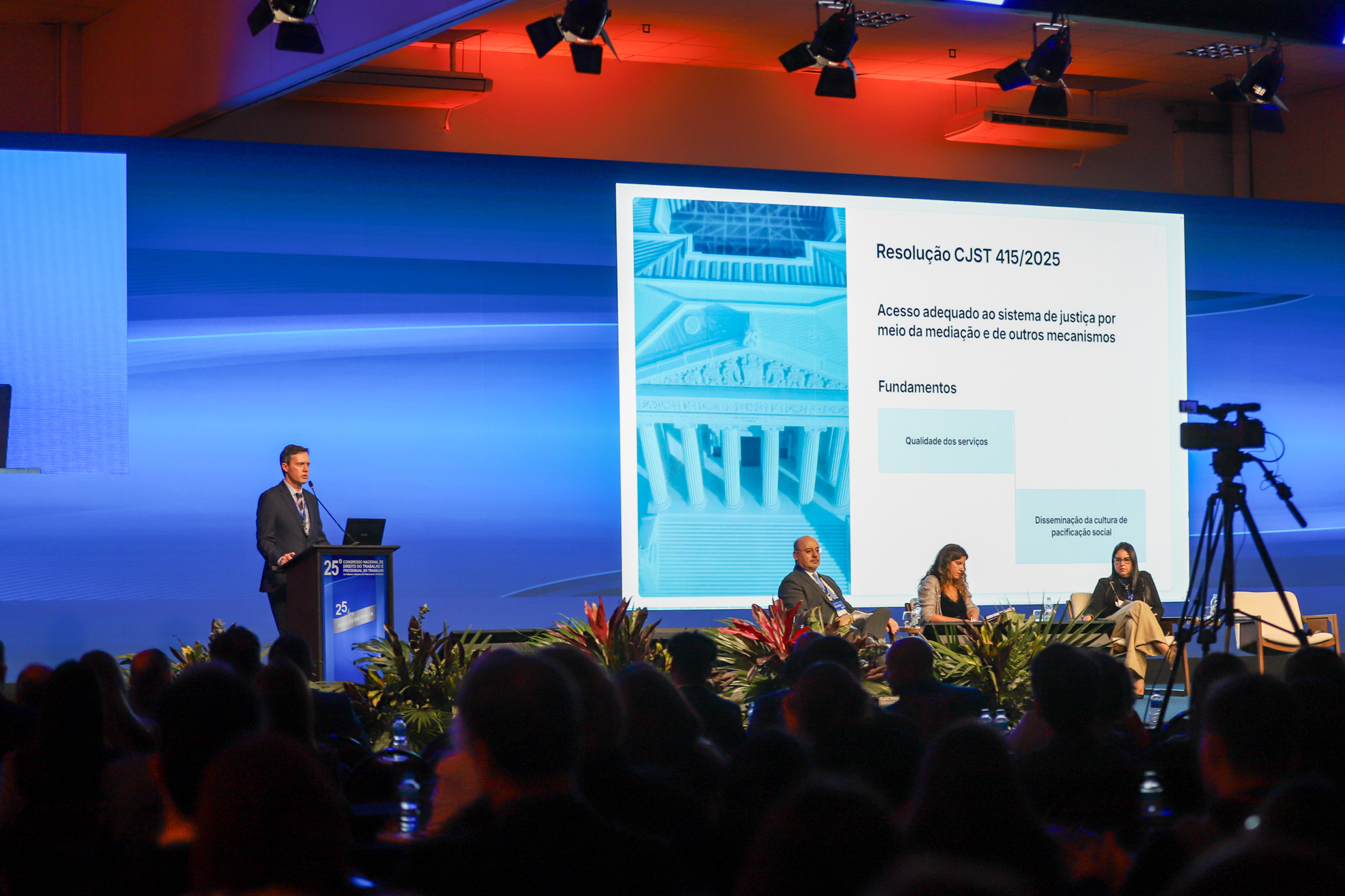 Auditório lotado durante o 25º Congresso Nacional de Direito do Trabalho e Processual do Trabalho. No palco, um homem de terno fala em um púlpito, enquanto um telão exibe a “Resolução CJST 415/2025” sobre acesso à justiça por mediação. Três pessoas estão sentadas à direita, e uma câmera grava o evento, com plantas decorando a frente do palco
