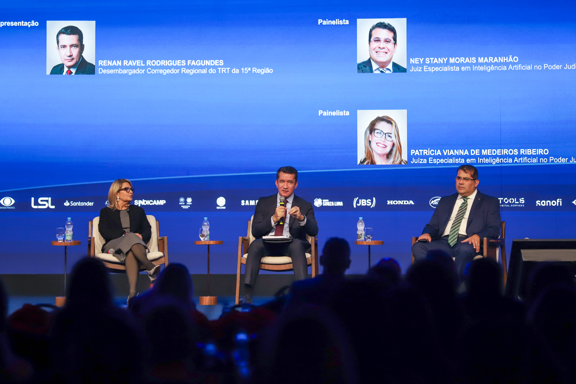 Três pessoas sentadas em poltronas no palco participam de um painel durante o 25º Congresso Nacional de Direito do Trabalho e Processual do Trabalho. Ao centro, um homem de terno escuro fala ao microfone; à esquerda, uma mulher de óculos e roupa preta e cinza ouve atentamente; e à direita, um homem de terno azul e gravata verde está sentado. No telão atrás deles, aparecem as fotos e nomes dos participantes: o mediador Renan Ravel Rodrigues Fagundes, desembargador corregedor regional do TRT da 15ª Região, e 
