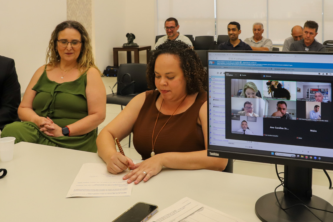 Mulher assina um papel que está sobre a mesa, sendo observada por outra, e com trabalhadores ao fundo, sentados. No lado direito da tela, um monitor com pessoas on-line