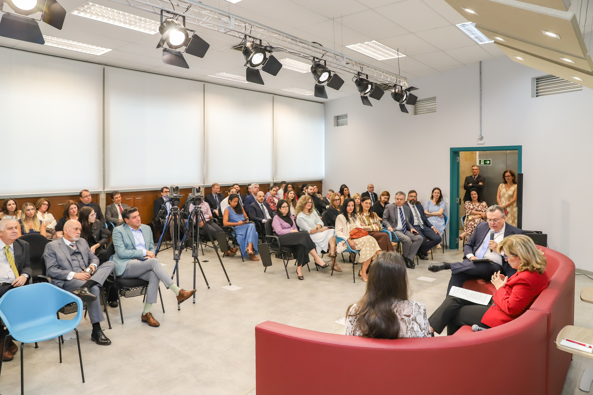 foto geral do auditório com foco no público, à direita da tela, os palestrantes estão sentados em um sofá vermelho
