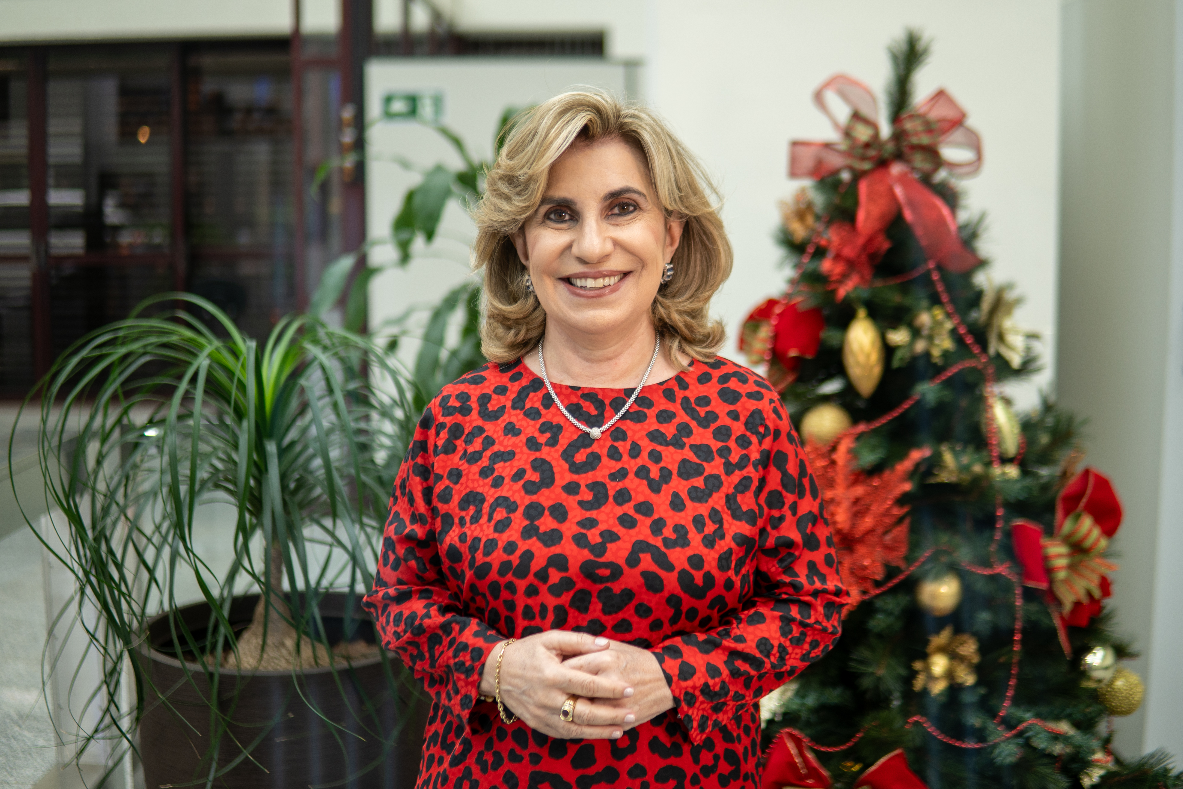 Dra. Ana Paula está de vermelho, num plano americano com fundo desfocado, em frente a uma árvore de natal, com mãos entrelaçadas