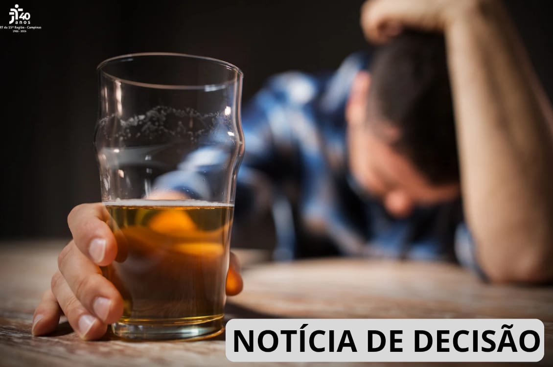 Em primeiro plano, um copo de bebida alcoólica sobre a mesa; ao fundo, uma pessoa desfocada apoia a cabeça no braço