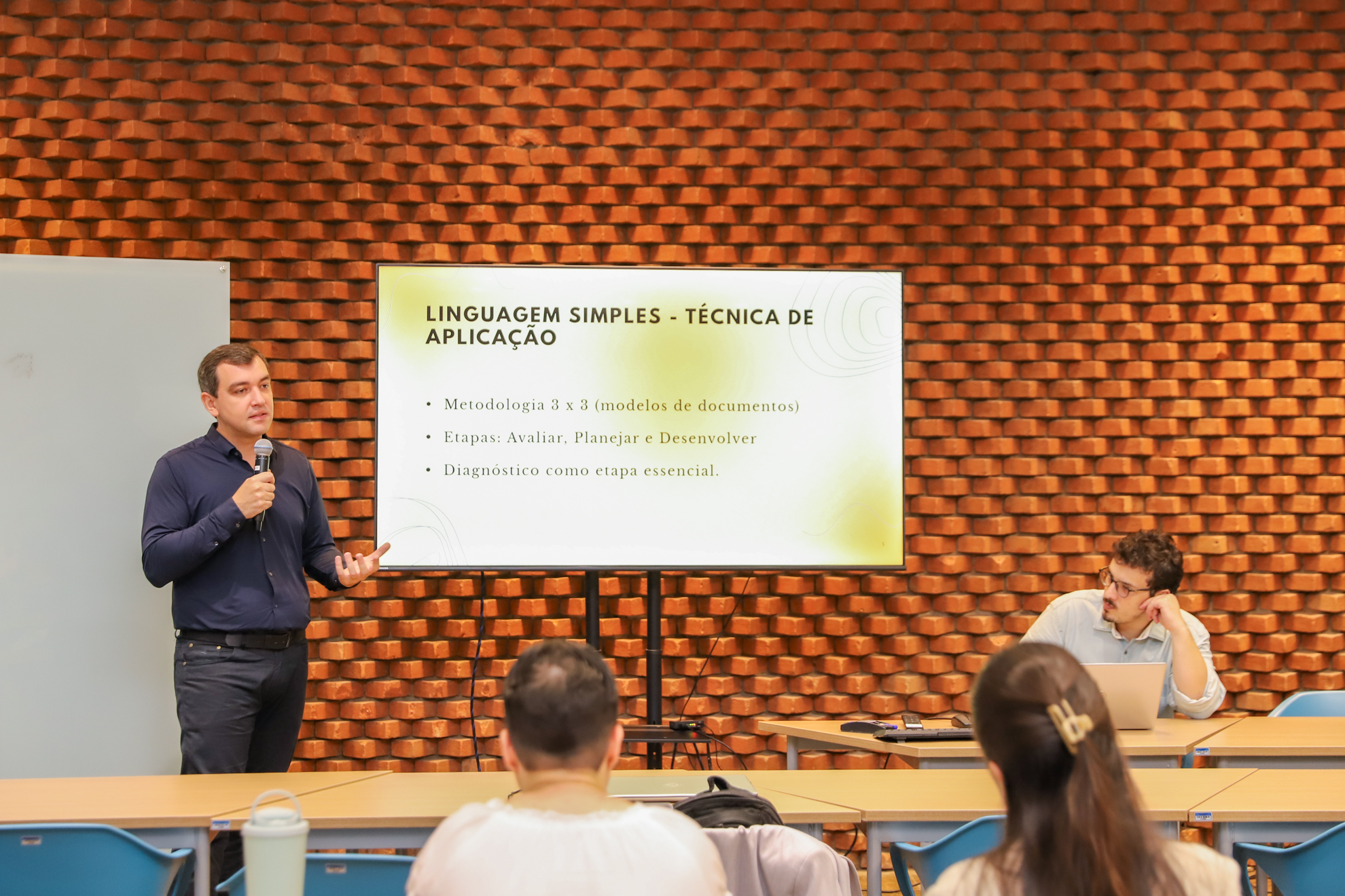 Thiago Rosa da Educorp está de pé com microfone na mão ao lado de uma tela que traz informações sobre linguagem simples. Em primeiro plano, os participantes do encontro acompanham a explanação