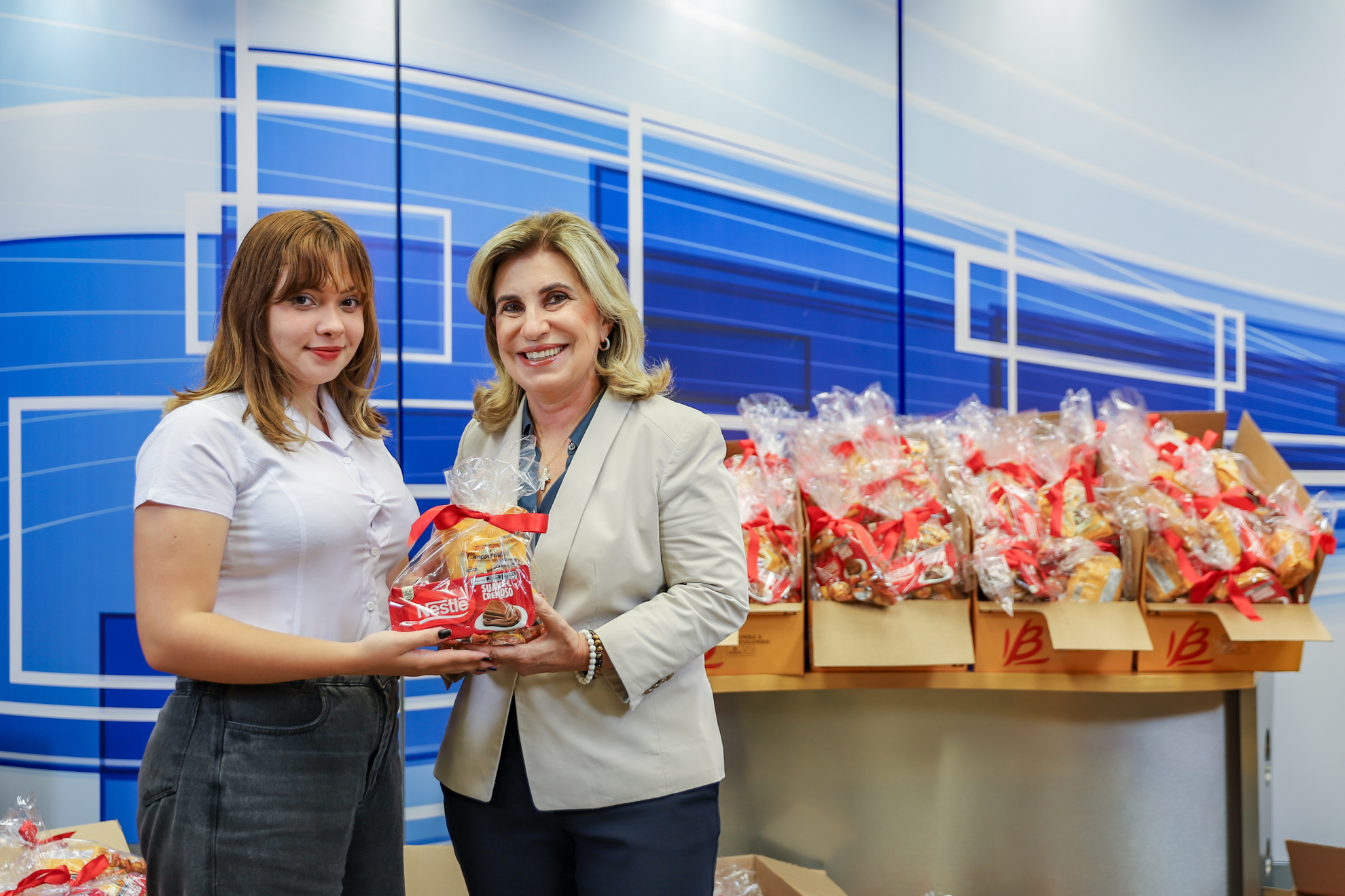 Dra. Ana Paula faz entrega do kit para uma moça, em frente a um painel azul e vários kits sobre a mesa