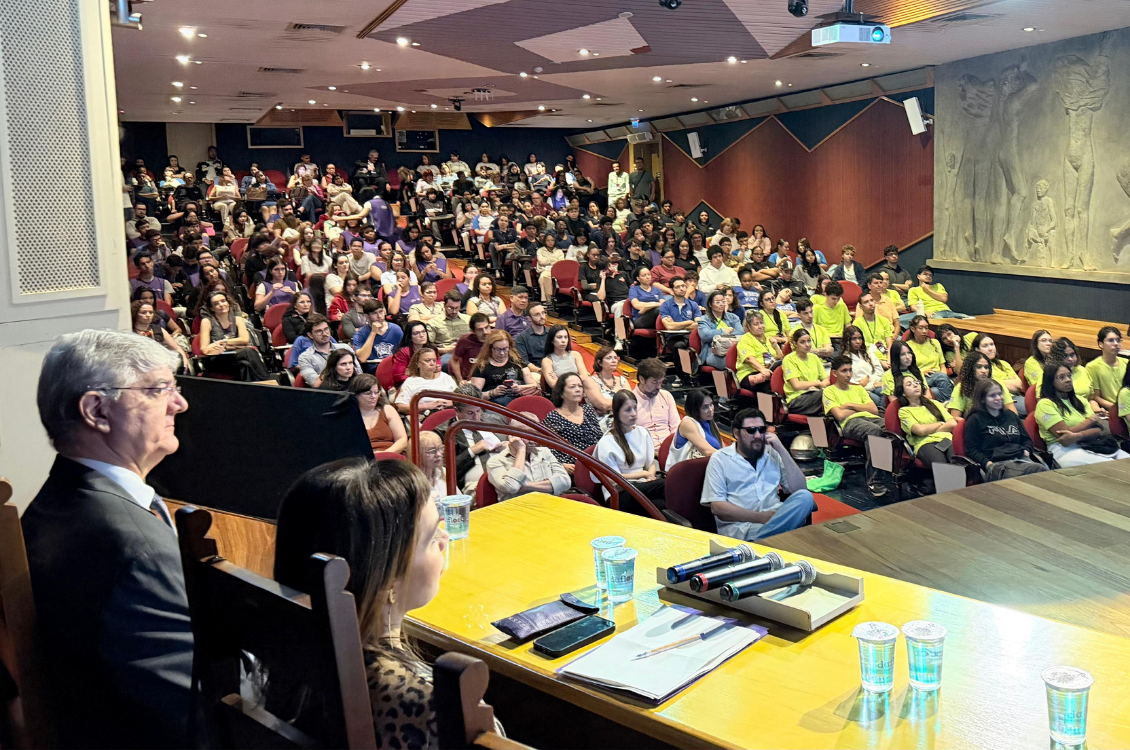plateia ao fundo com o Dr. Pelegrini e Dra. Marina em primeiro plano, do lado esquerdo, sentados à mesa