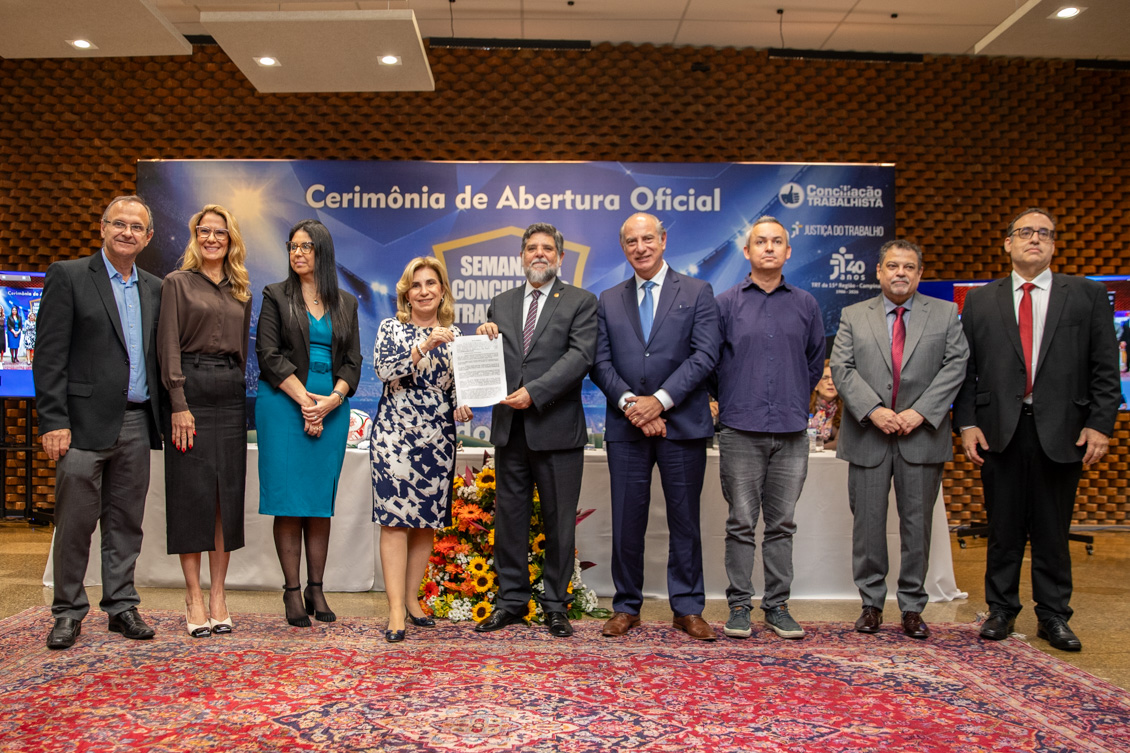Assinantes do acordo estão de pé em frente ao banner alusivo ao evento, com a Dra. Ana Paula e o ministro Caputo Bastos exibindo o acordo assinado