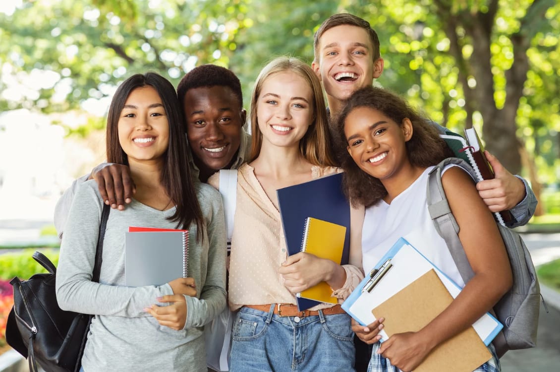 cinco estudantes sorridentes, de várias etnias, segurando livros e cadernos em um local aberto, com árvores