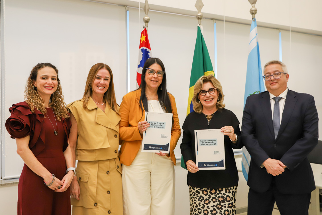 "Foto colorida, em ambiente institucional. Cinco pessoas estão lado a lado, sendo quatro mulheres e um homem, todos em pé e sorrindo para a câmera. Duas das mulheres, posicionadas ao centro, seguram exemplares de uma publicação. Ao fundo, há bandeiras em mastros, incluindo a do Brasil e outras bandeiras institucionais, além de grandes janelas com persianas claras. A imagem transmite um momento de registro oficial de entrega ou lançamento de material."