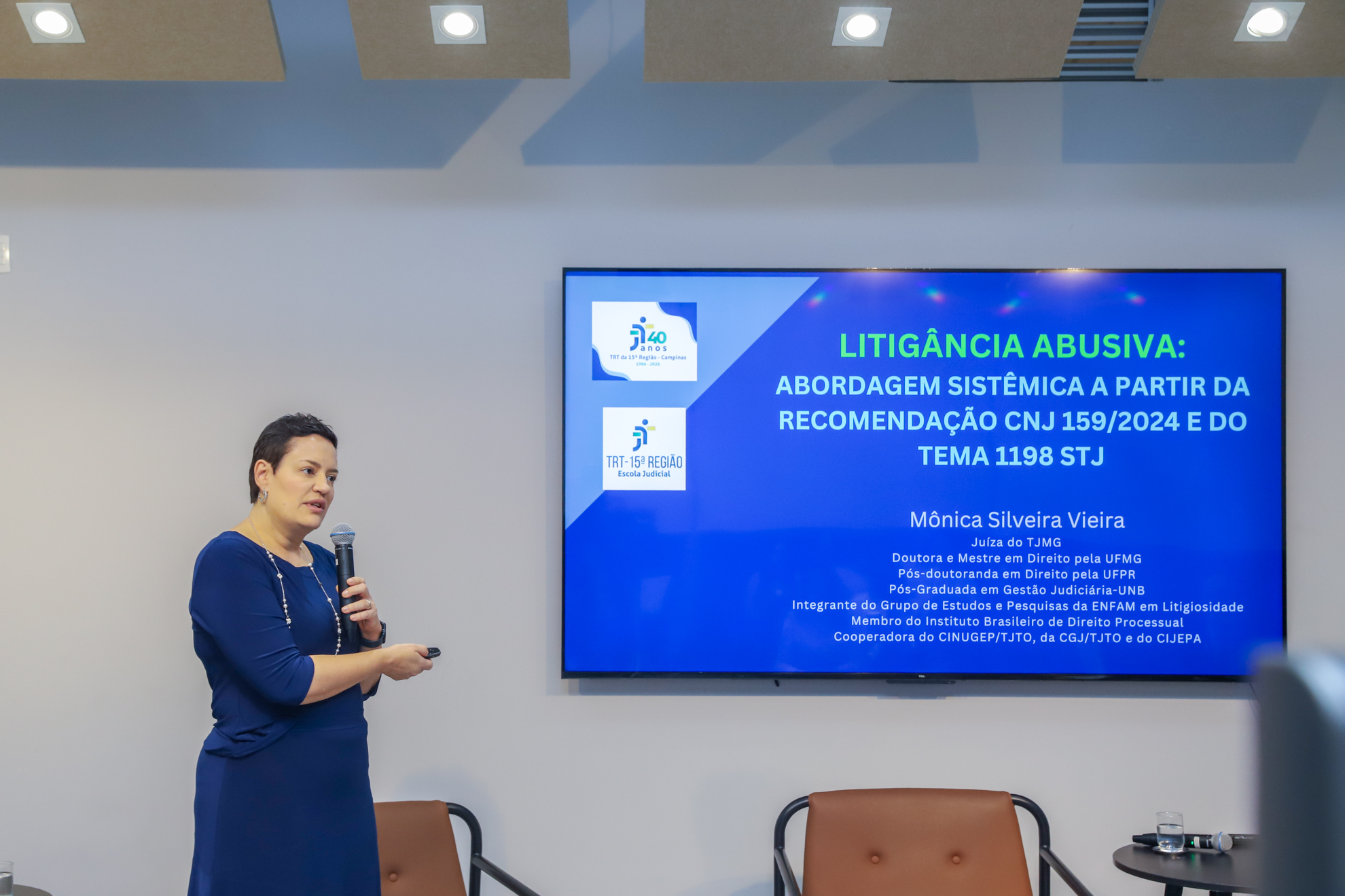 juíza Mônica fala ao microfone de pé, apontando para uma tela com dados sobre o tema