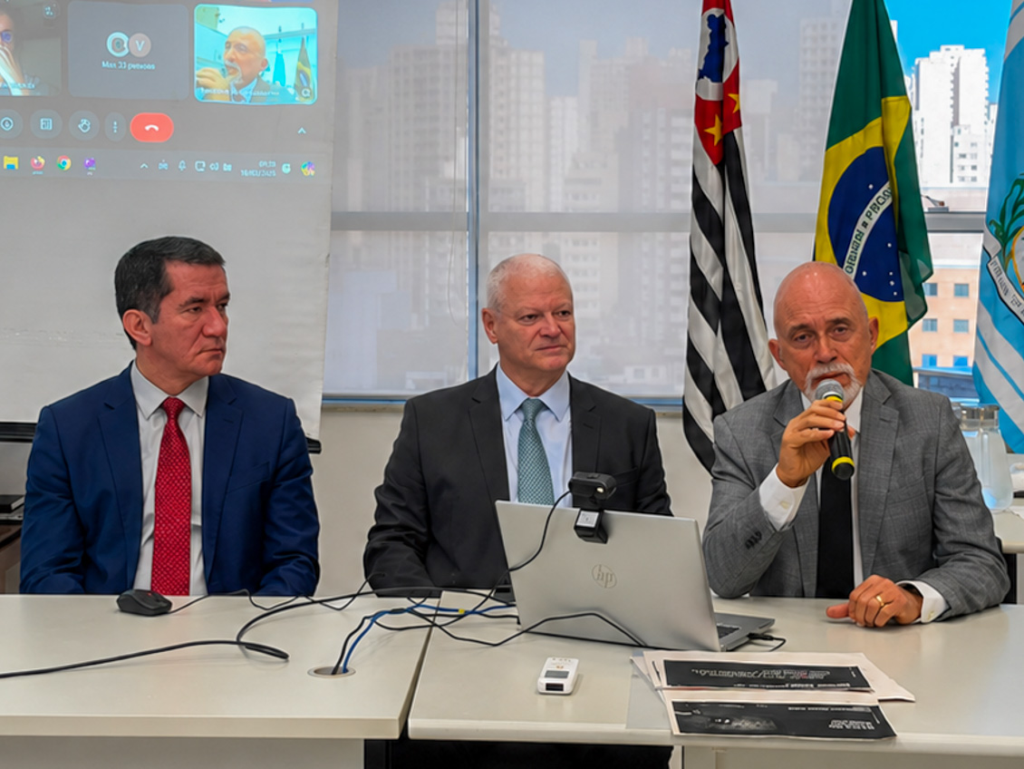 Três homens sentados, um deles fala ao microfone