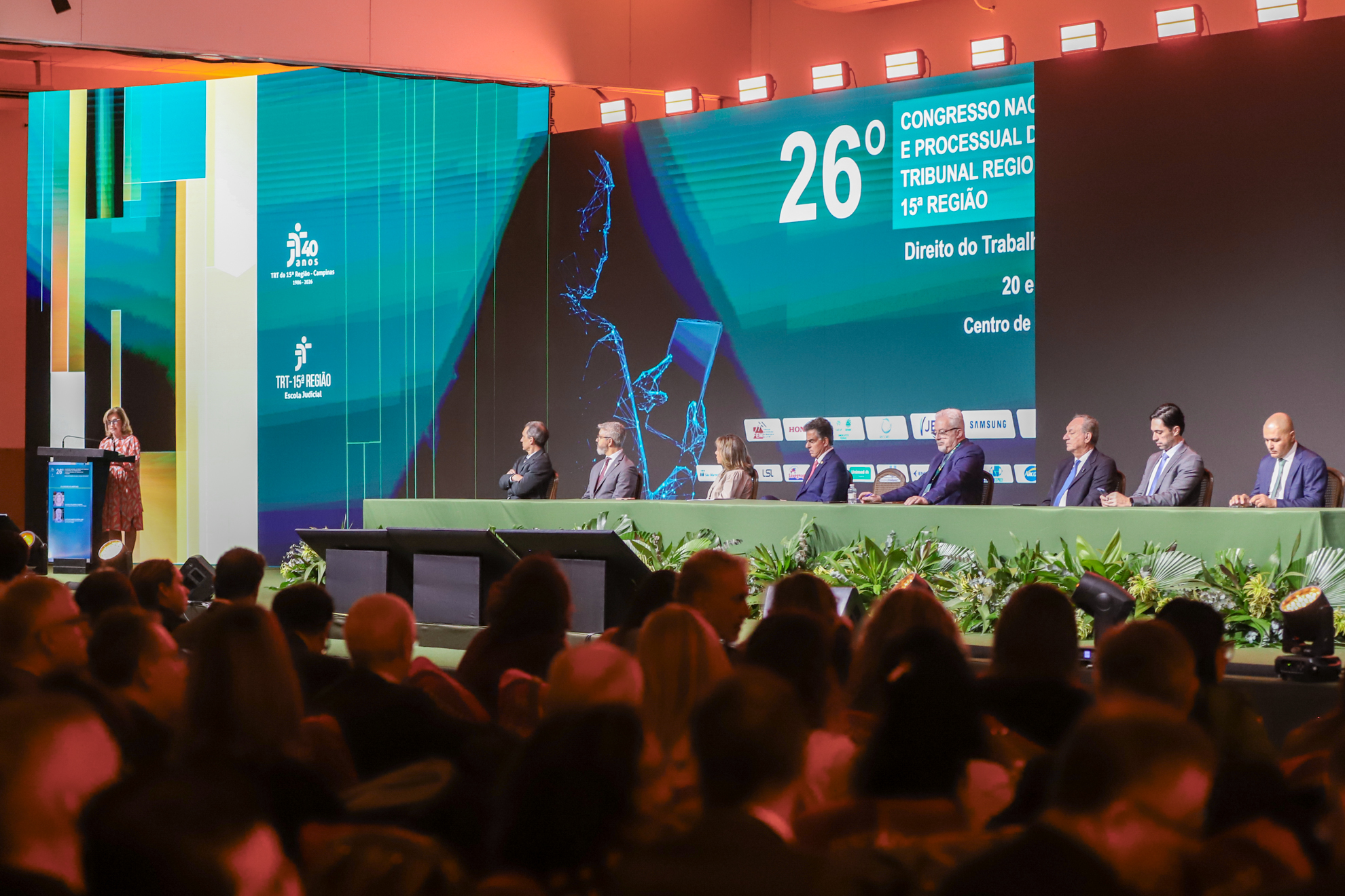 A imagem mostra um grande auditório durante um congresso jurídico, visto a partir da plateia.  No palco, há uma longa mesa revestida de verde, decorada na parte frontal com arranjos de plantas. Oito pessoas estão sentadas atrás da mesa, aparentemente compondo uma mesa de autoridades ou palestrantes. À esquerda do palco, uma mulher está em pé diante de um púlpito, aparentemente falando ao público.  Ao fundo, grandes painéis de LED exibem uma identidade visual em tons de azul, turquesa e verde, com elementos 