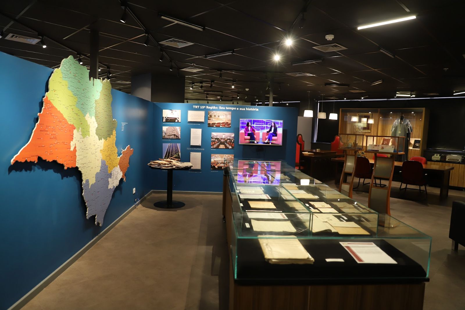 Ao lado esquerdo há um mapa do Estado de São Paulo pregado em uma parede azul. Ao centro e em primeiro plano, uma mesa de vidro com base de madeira, com documentos antigos dentro. Ao fundo da mesa, dispostos em uma parede de cor azul, há seis fotografia e três documentos escritos. Ao lado direito, na mesma parede um TV passa imagens. Do lado direito, uma mesa de madeira escura com cadeiras vermelhas. Ao lado direito da mesa, em um armário com porta de vidro, está disposto uma veste talar, preta e no teto, três lustres iluminam a mesa.s e 