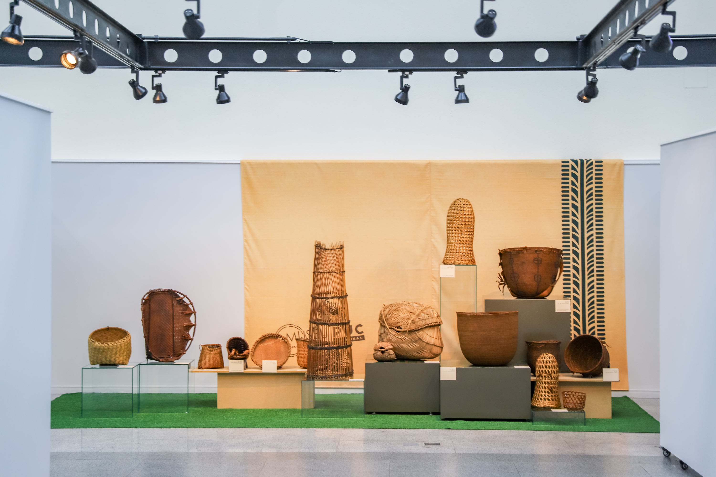"objetos indígenas em palha expostos em totens de acrílico e mesa de madeira"