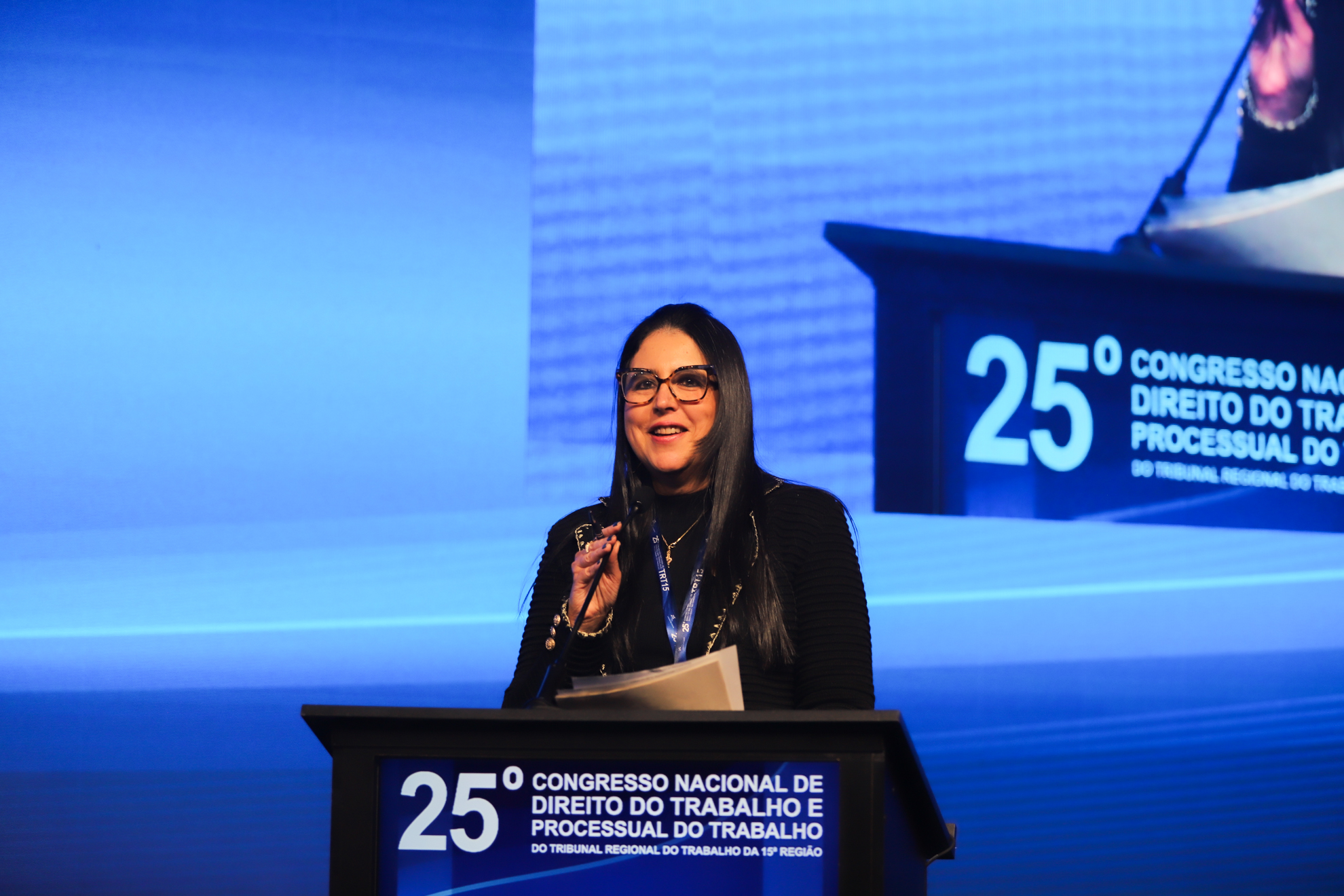 "Mulher de cabelos longos e lisos, usando óculos, blusa preta e crachá, fala sorrindo em um púlpito durante o 25º Congresso Nacional de Direito do Trabalho e Processual do Trabalho. Ela segura papéis na mão esquerda, e ao fundo há um telão que projeta sua imagem ampliada"