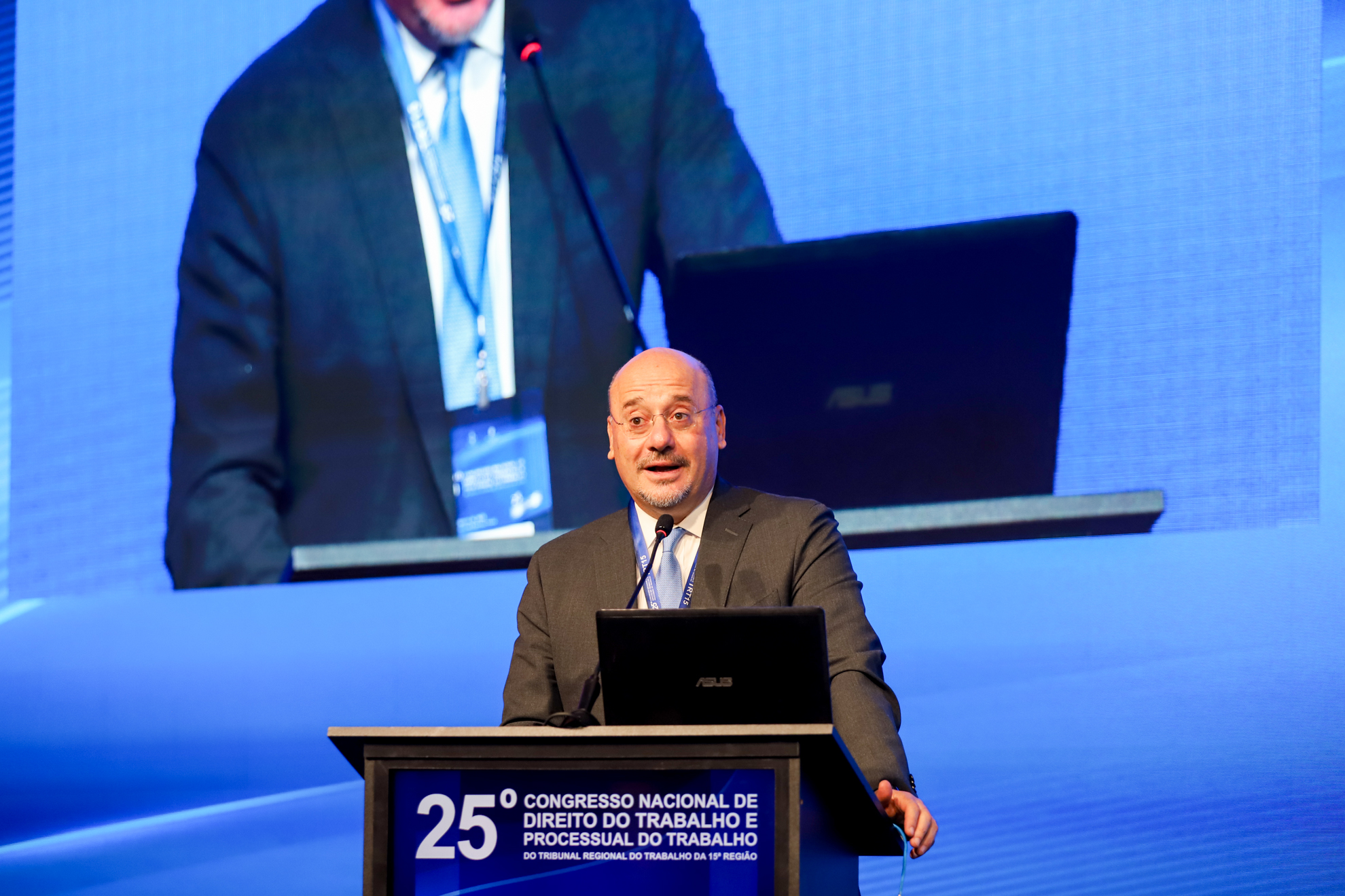 "Homem de óculos e terno escuro com gravata azul-clara, fala em um púlpito durante o 25º Congresso Nacional de Direito do Trabalho e Processual do Trabalho. Atrás dele, um telão exibe sua imagem ampliada, e um notebook está aberto sobre o púlpito"