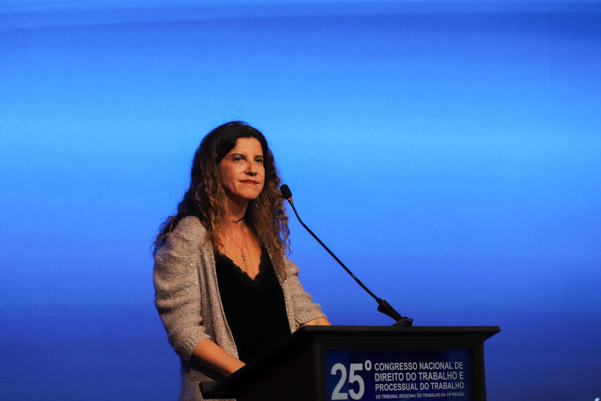 "Mulher de cabelos castanhos ondulados, usando colar e blusa preta com casaquinho prateado, fala em um púlpito durante o 25º Congresso Nacional de Direito do Trabalho e Processual do Trabalho. O fundo é iluminado em azul, e o microfone está posicionado à sua frente"