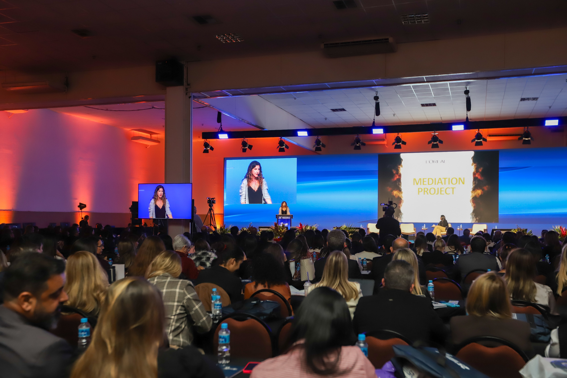 "Auditório cheio durante o 25º Congresso Nacional de Direito do Trabalho e Processual do Trabalho. No palco, uma mulher fala em um púlpito, enquanto sua imagem é projetada em dois telões. Ao fundo, um grande painel exibe o título “Mediation Project” com a logomarca da L’Oréal. A plateia, formada por dezenas de pessoas sentadas, acompanha a apresentação, e câmeras registram o evento. As paredes estão iluminadas com luzes laranja e o cenário de fundo é azul."