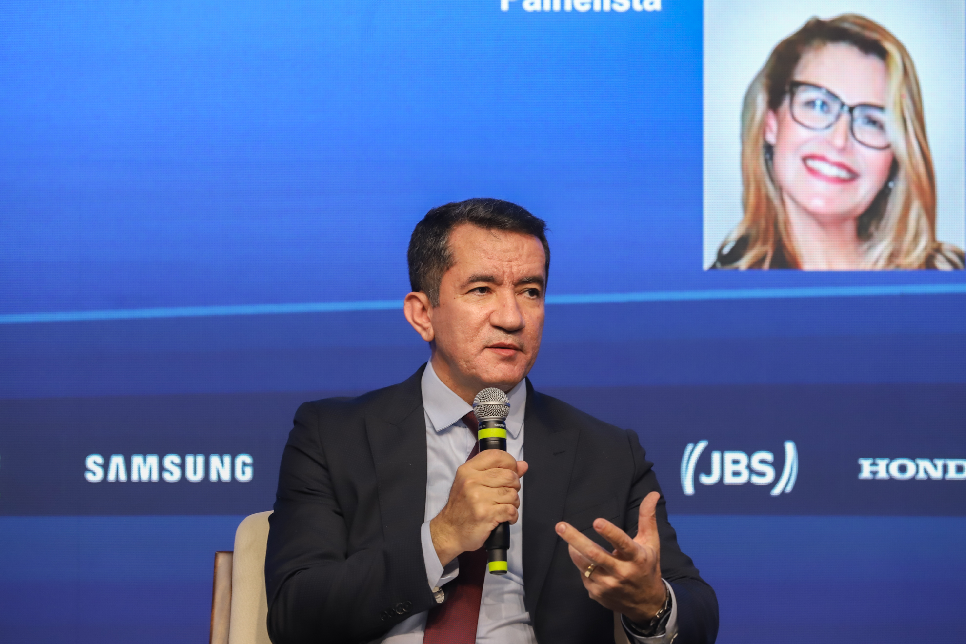"O desembargador Renan Ravel Rodrigues Fagundes, corregedor regional do TRT da 15ª Região, fala ao microfone durante o 25º Congresso Nacional de Direito do Trabalho e Processual do Trabalho. Ele veste terno escuro com camisa azul-clara e gravata vinho, gesticulando com a mão esquerda enquanto responde ou comenta. Ao fundo, o telão exibe parte da foto e nome de outra participante do painel"