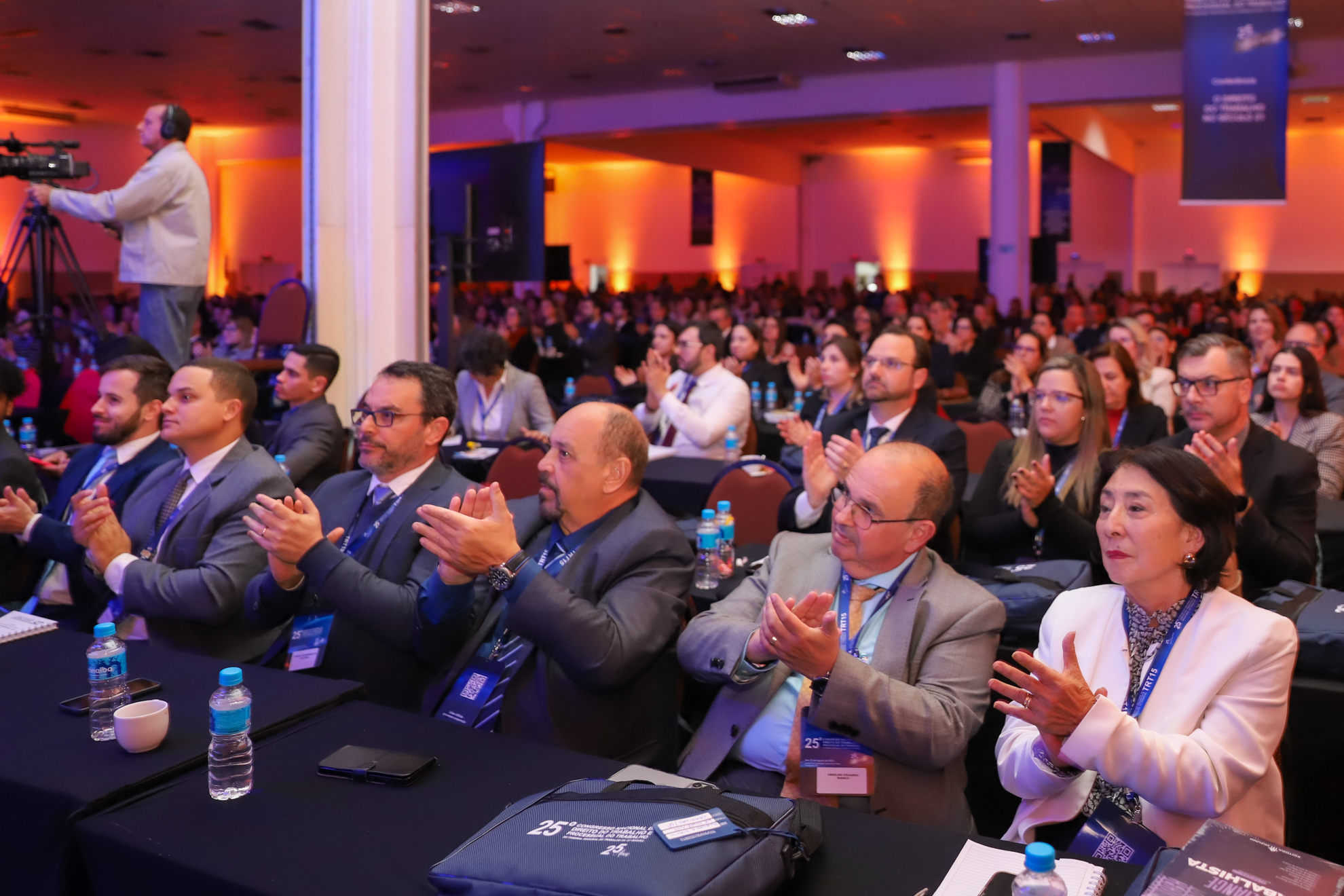 "Plateia do 25º Congresso Nacional de Direito do Trabalho e Processual do Trabalho aplaude durante uma apresentação. Na frente, um grupo de homens e mulheres, muitos usando crachás do evento e trajes formais, está sentado em mesas com garrafas de água e materiais de anotação. Ao fundo, o auditório está lotado e iluminado com luzes em tons de laranja, enquanto um cinegrafista à esquerda registra a cena com uma câmera profissional"