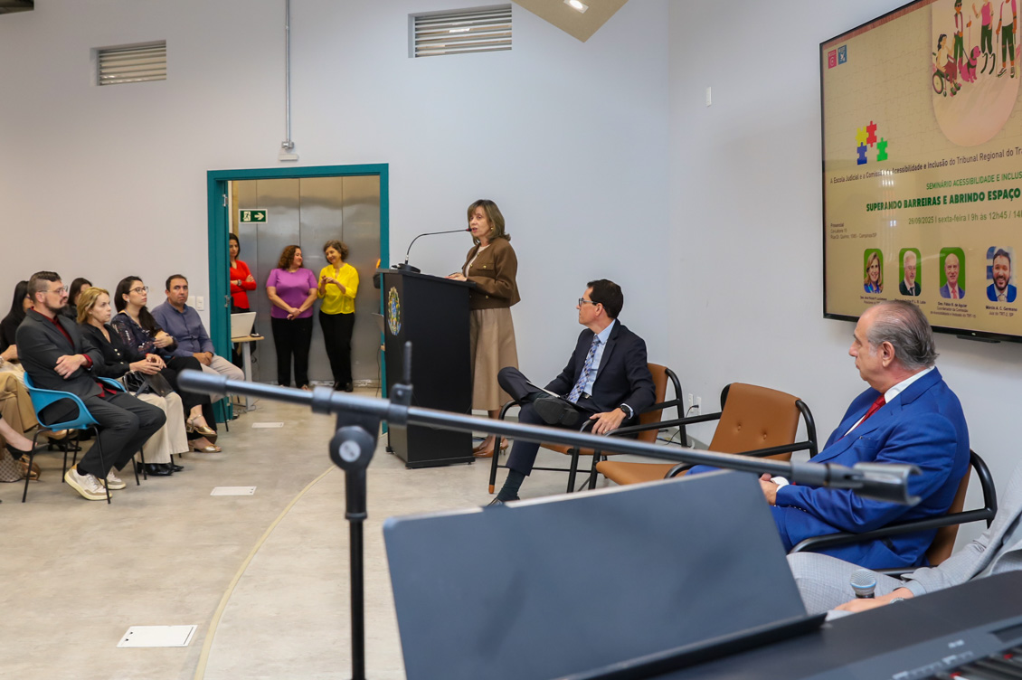 "procuradora Alvamari está de pé no púlpito, falando ao microfone, sendo observada pelo público e pelos integrantes da mesa alta"