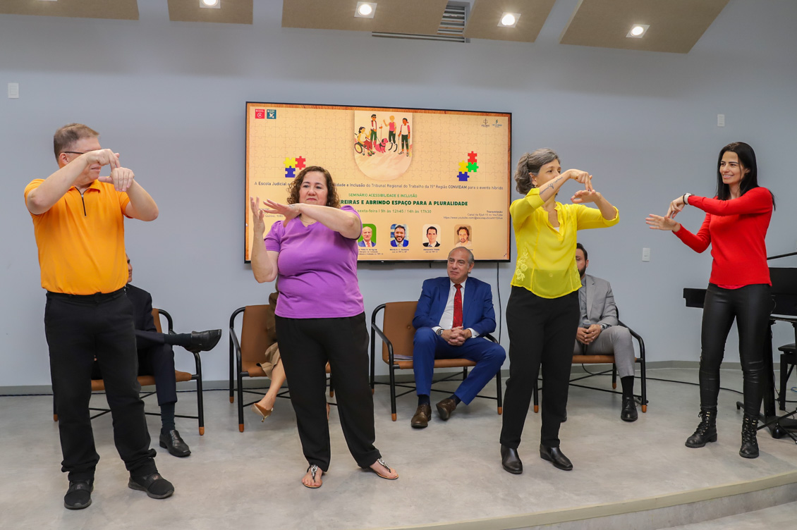 "Quatro integrantes do Coral de Libras apresentam música, com integrantes da mesa alta ao fundo"