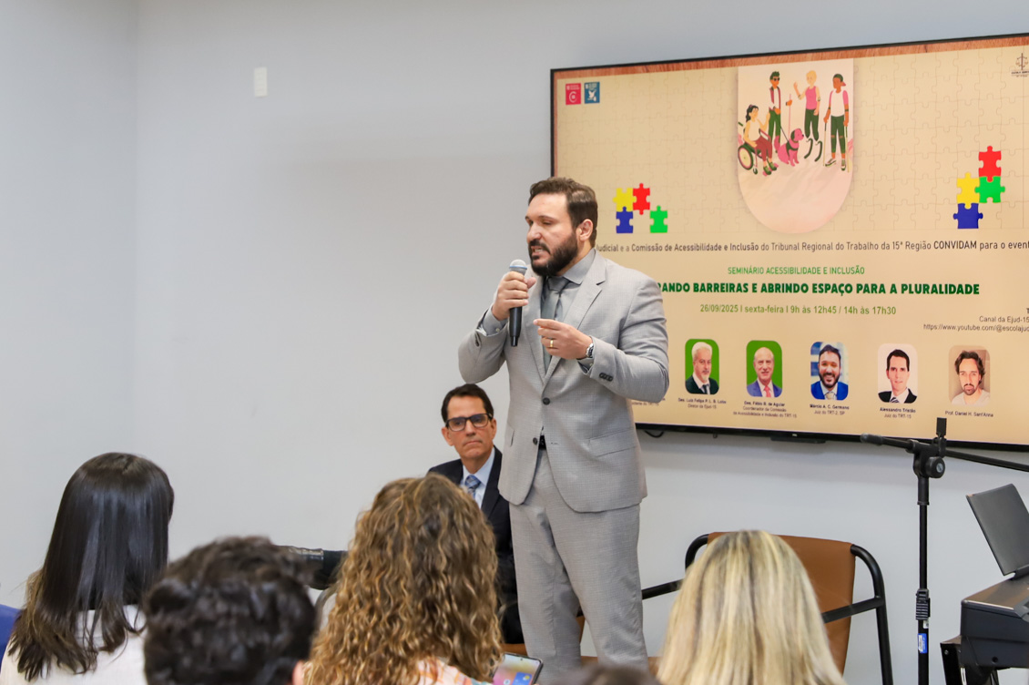 "Juiz Márcio está de pé, falando ao microfone, sendo observado pelo juiz Tristão e pelo público que lota o auditório"