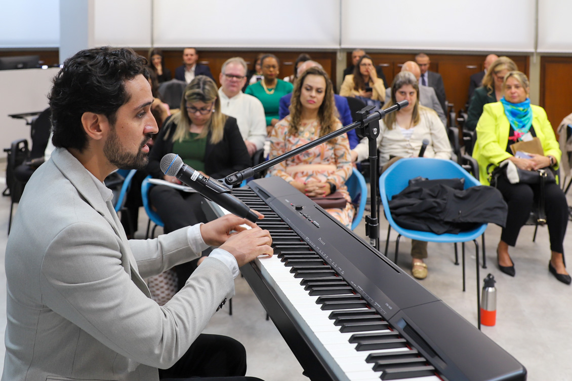 "Professor Daniel está diante de um teclado, sendo observado pelo público ao fundo"