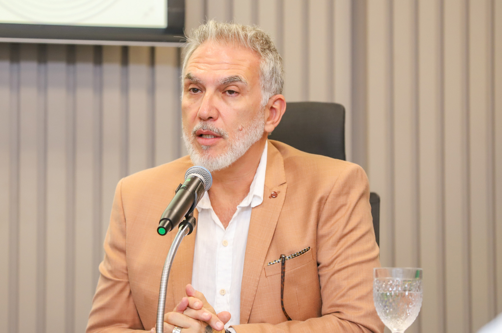 "Dr. Carlos Eduardo está sentado à mesa alta, falando ao microfone"