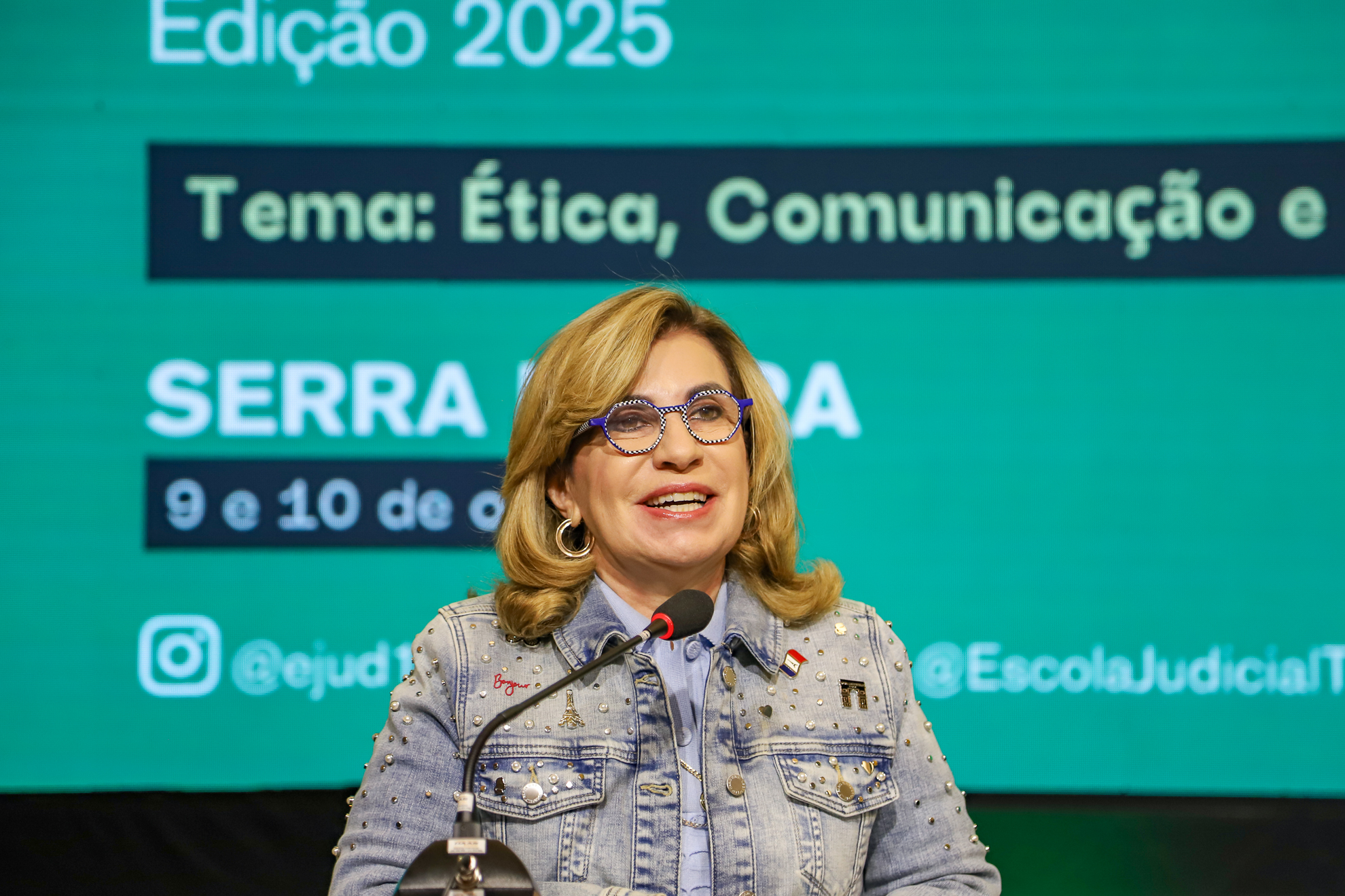 "Dra. Ana Paula fala ao microfone. Ao fundo painel verde, com texto em branco sobre retângulo azul, contendo dados sobre a temática do evento"