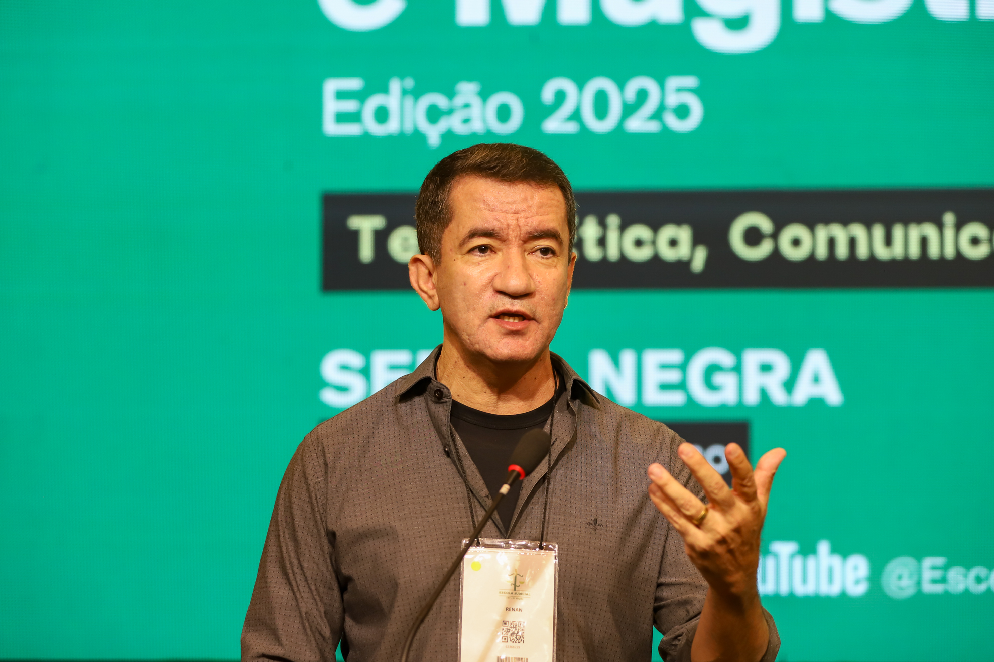 "Dr. Fenan fala ao microfone. Ao fundo painel verde, com texto em branco sobre retângulo azul, desfocado"