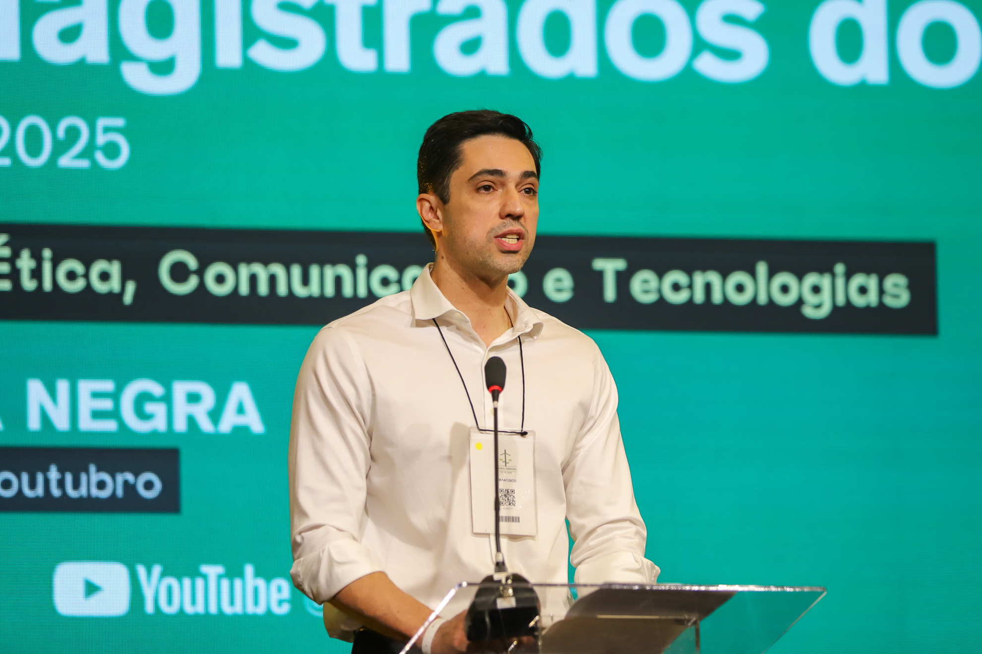 "Dr. Francisco fala ao microfone. Ao fundo painel verde, com texto em branco sobre retângulo azul, desfocado"