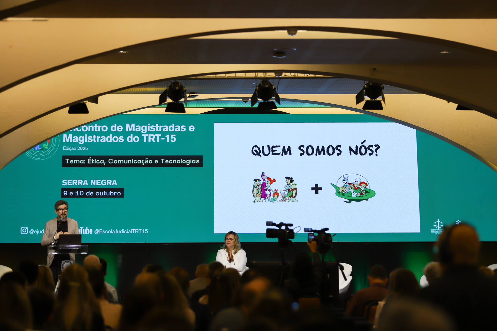 "mesa alta com o psicólogo Alessandro Marimpietri falando ao microfone, sendo observado pela Dra. Eleonora. Participantes acompanham sentados no auditório."