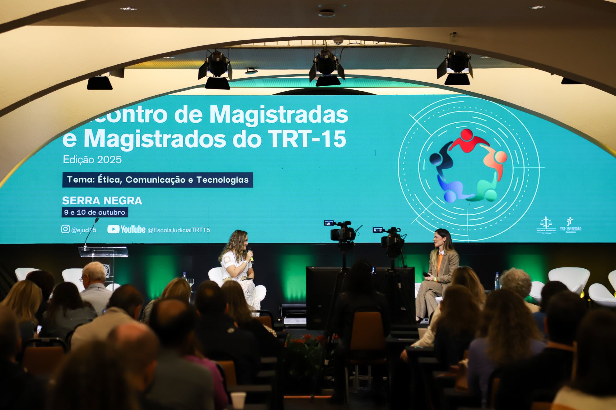 "Duas mulheres estão lado a lado no palco, com microfone nas mãos, sendo observadas pelo público no auditório"