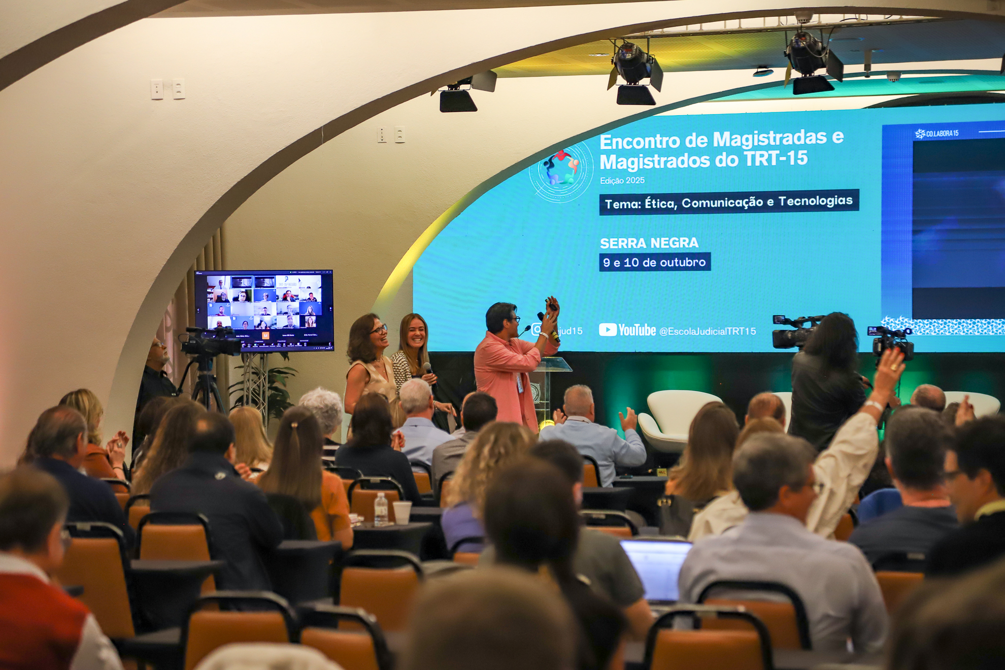 "Três integrantes do Colabora estão ao fundo, sendo observados pela plateia. Na TV, magistrados acompanham o evento no formato on-line"